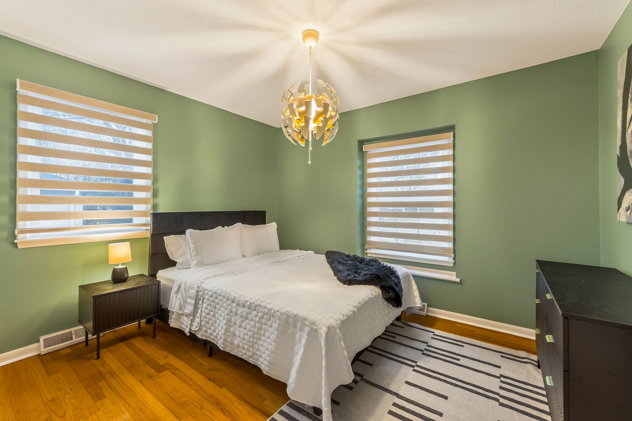A bedroom with light green walls, hardwood floors, and two striped window blinds. The bed has white bedding and pillows, with a dark headboard. There is a black nightstand with a lamp on it, and a black dresser on the right. A modern chandelier hangs