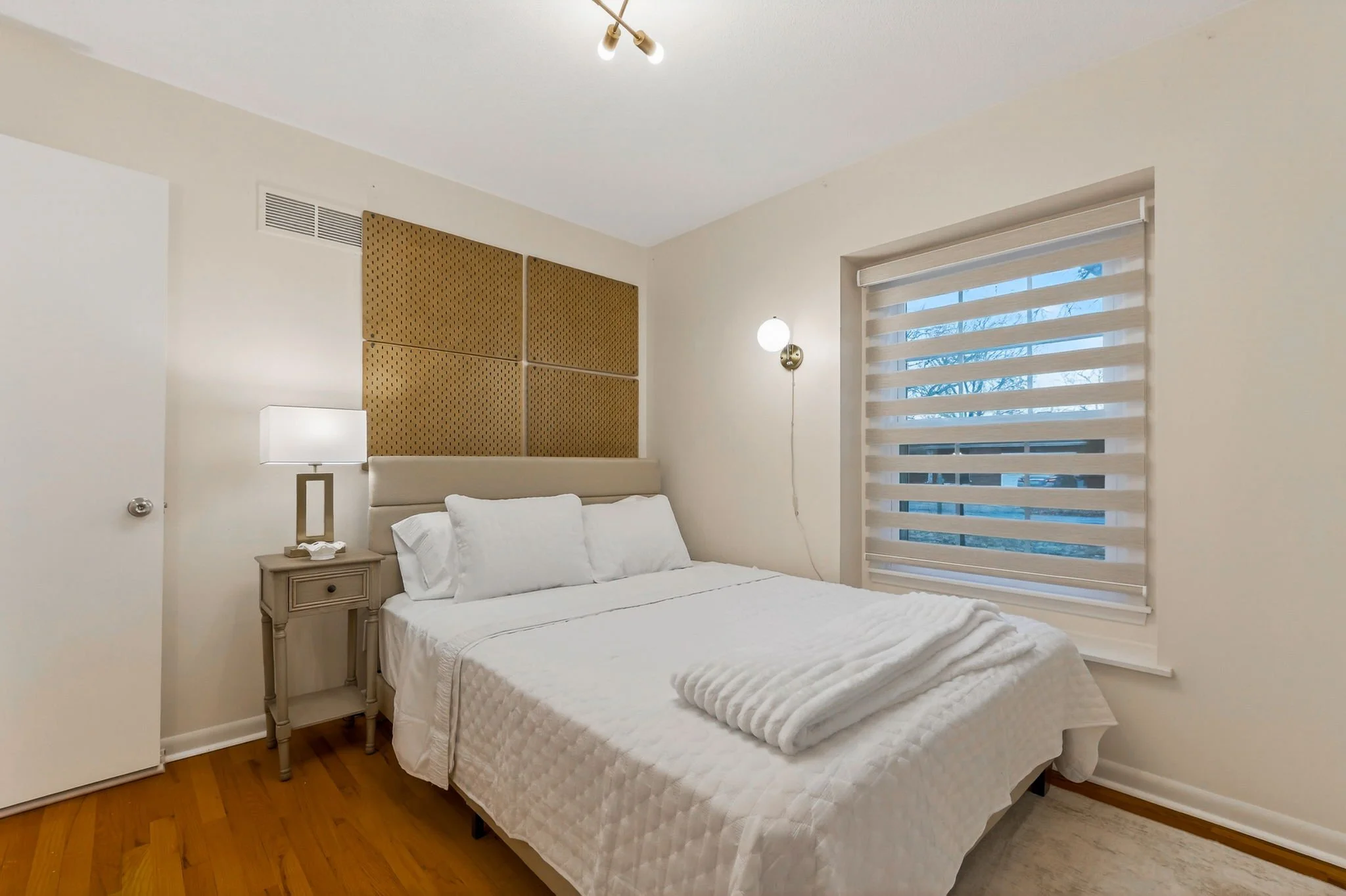 A bedroom with a white bed, beige and gold decor, a side table with a white lamp, a window with horizontal blinds, and a modern ceiling light fixture.