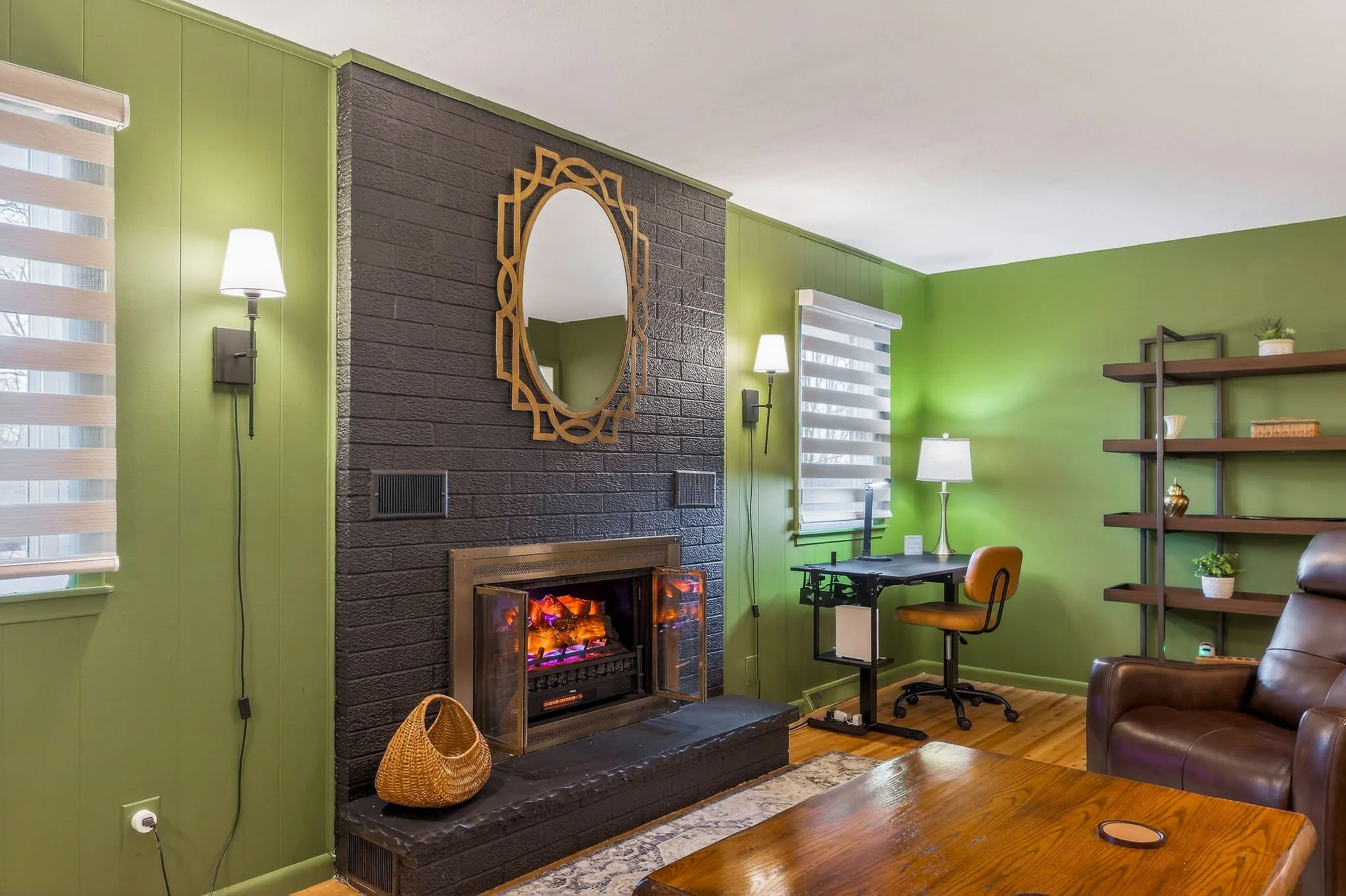 Living room with green walls, a black brick fireplace with a mirror above, a wooden coffee table, a leather armchair, a small desk with a yellow chair, a bookshelf, and various lamps, with hardwood flooring.
