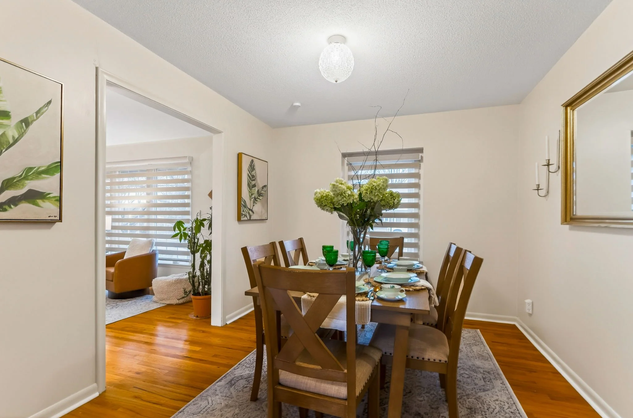 Dining room with wooden table, six chairs, green glasses, and a floral centerpiece, with wall art and a mirror on the walls, and hardwood flooring.