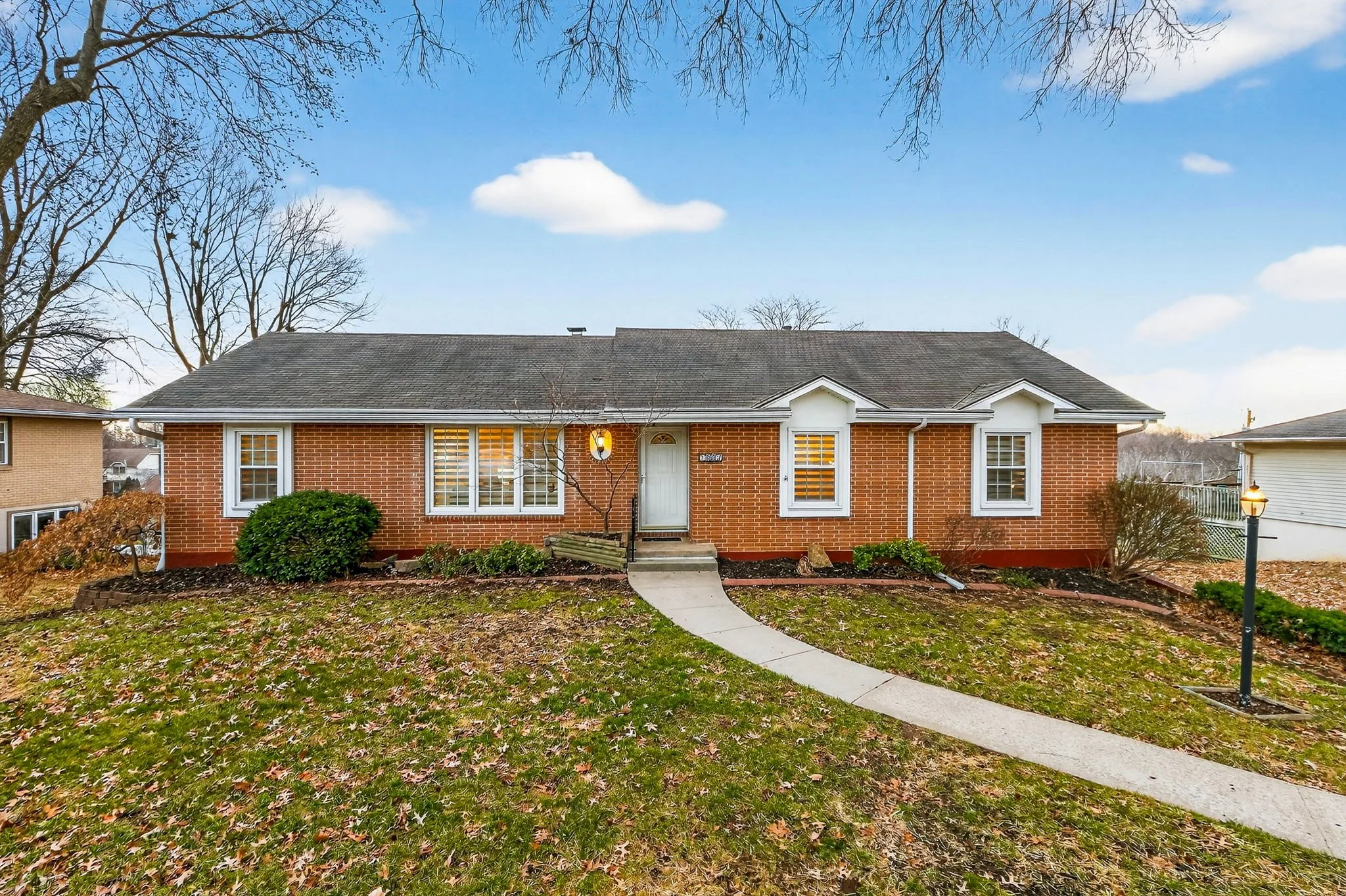 Single-story brick house with a gabled roof, front yard path, shrubs, and leafless trees under a partly cloudy sky.