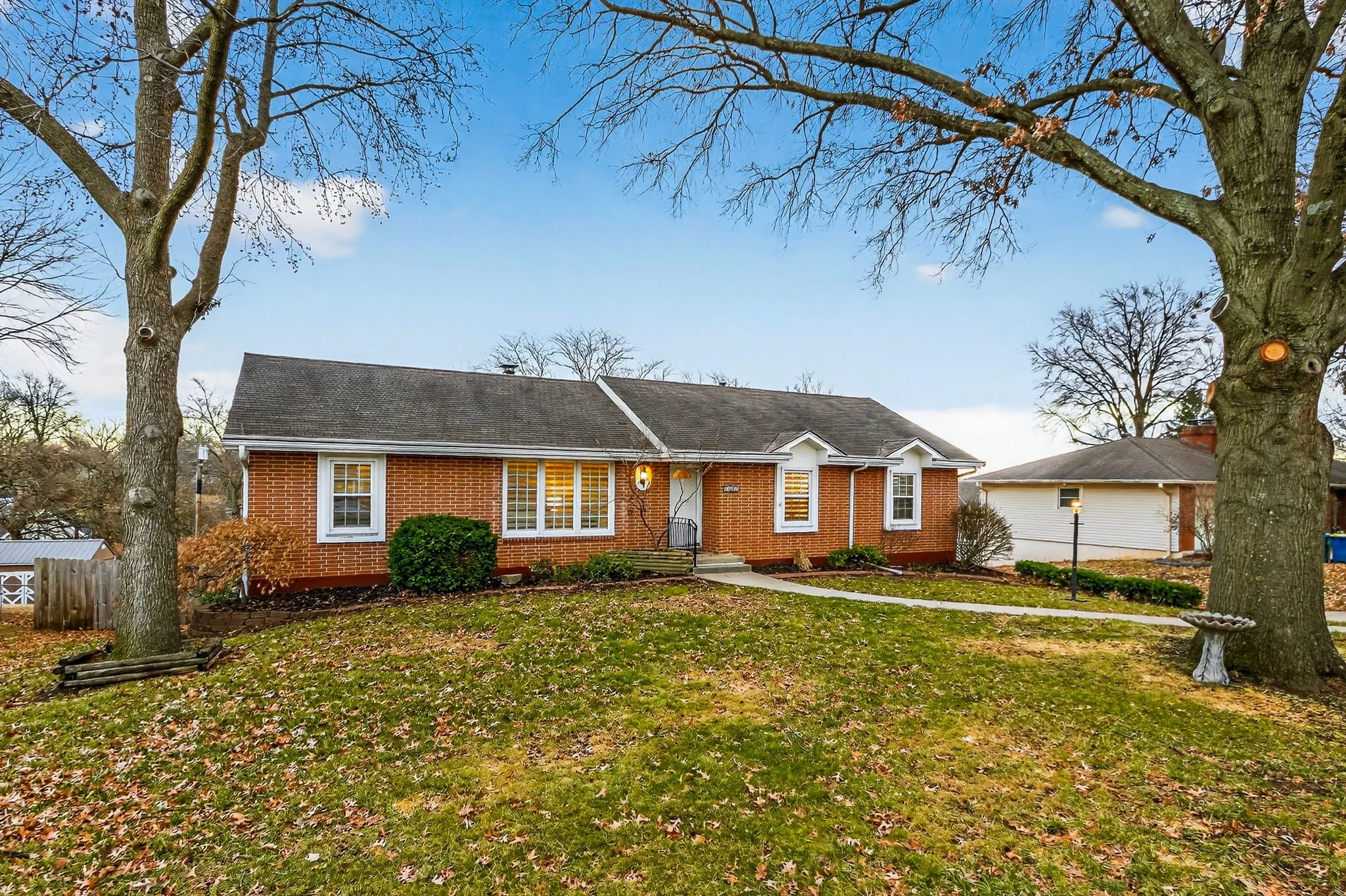 Front view of a brick house with large front yard. Two trees frame the scene, and the grass is covered with fallen leaves. The house has a small porch with steps and white window shutters. There are two small bushes near the front door and a pathway 