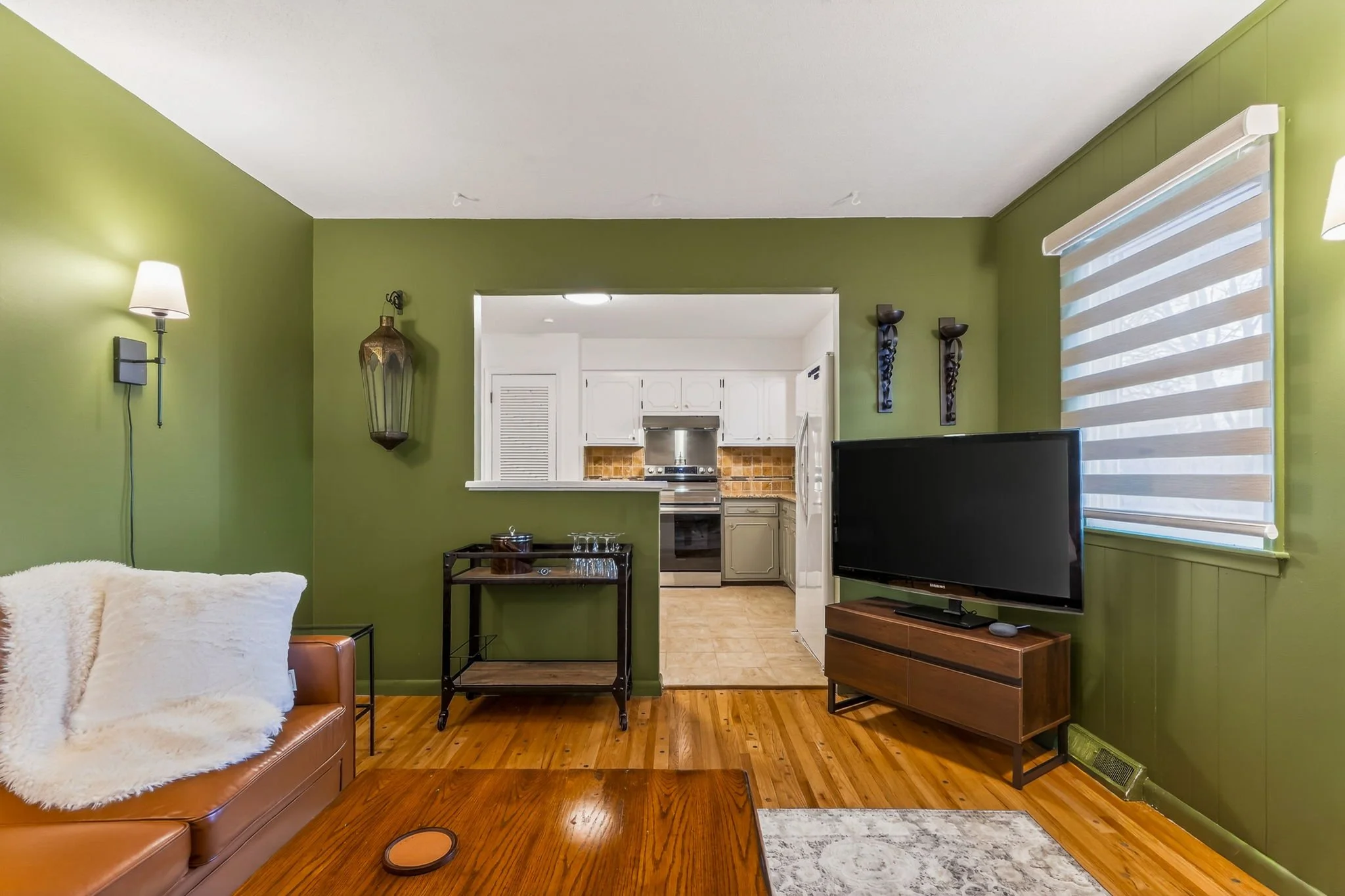 Living room with green walls, brown leather sofa with a white throw, wooden coffee table, flat-screen TV on a wooden stand, large window with striped blinds, view into a kitchen with white cabinets and stainless steel appliances.