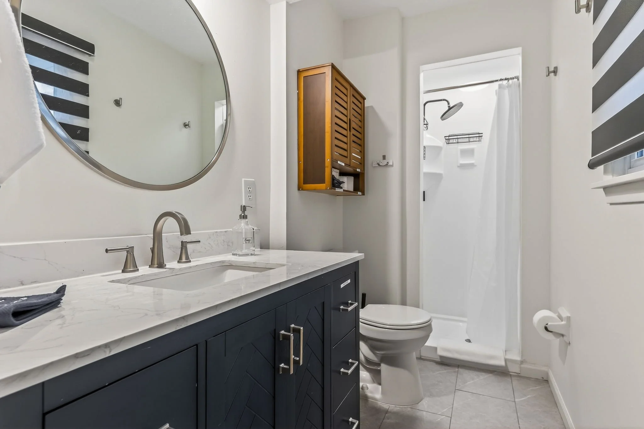 A bathroom with a navy blue vanity, marble countertop, and silver fixtures. A round mirror, a soap dispenser, and a black washcloth are on the counter. There is a toilet, a small wooden cabinet on the wall, and a shower with a white curtain and a gra