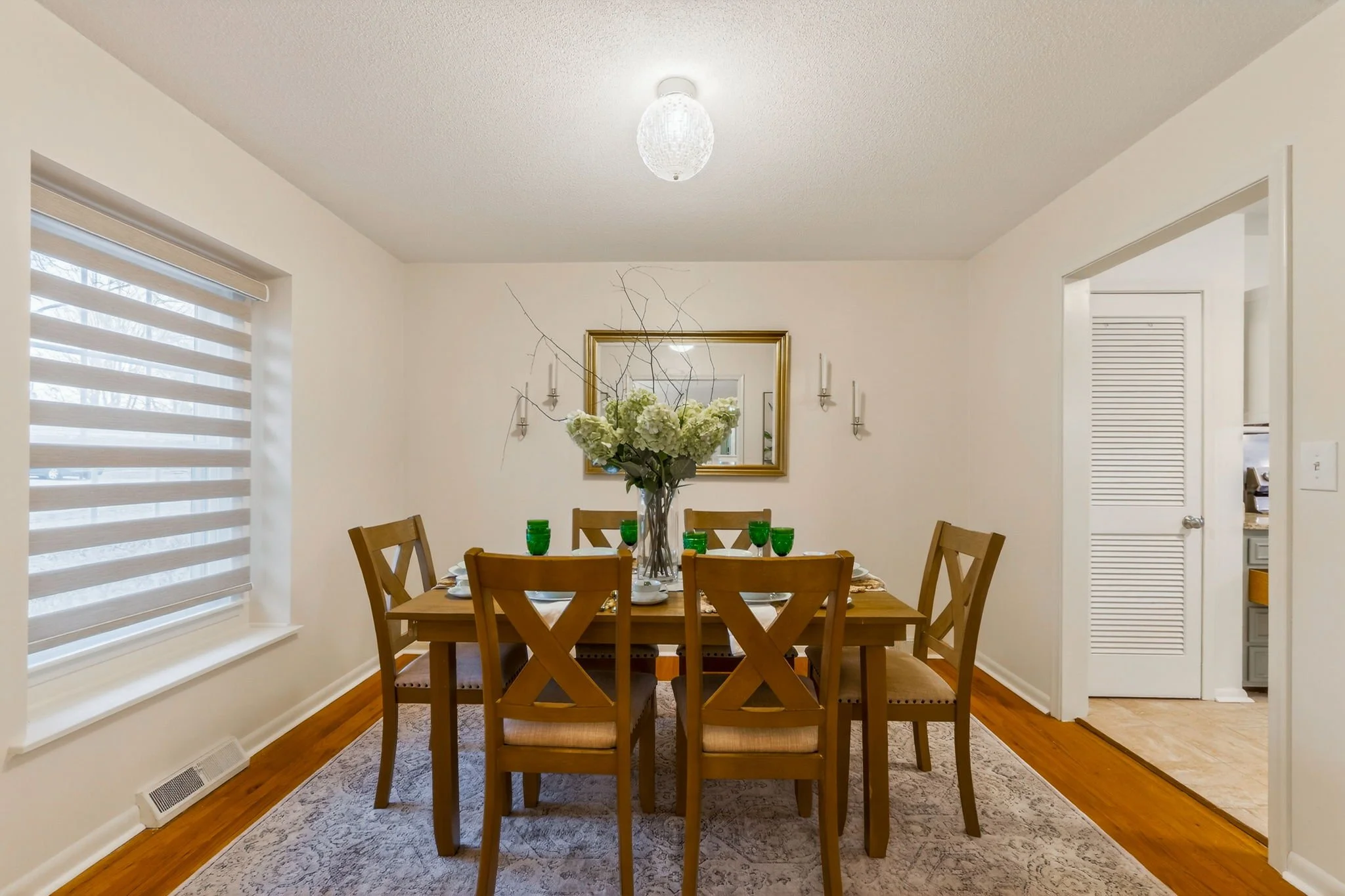 Dining room with a wooden table set with six green glasses, a large floral centerpiece, and six placemats. There is a mirror on the wall behind the table and wall-mounted candle sconces. The room has a window with horizontal blinds, hardwood flooring