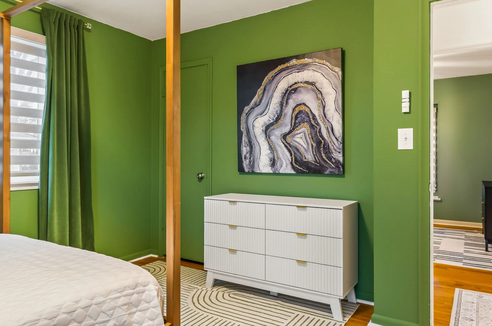 A bedroom corner with green walls, a white dresser beneath a framed abstract black, white, gray, and gold art piece, and a window with green curtains.