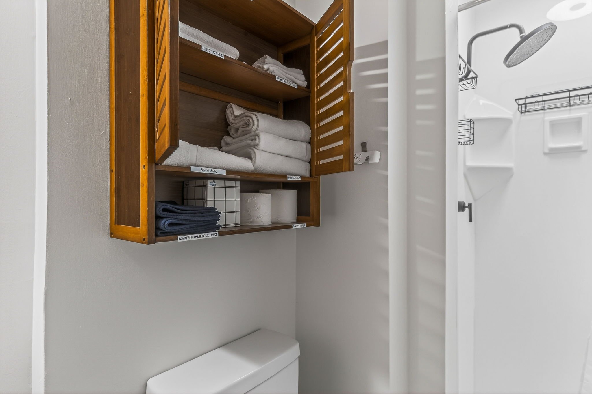 Bathroom with wooden cabinet containing folded towels, washcloths, and toilet paper, a toilet, and part of shower with a rain showerhead.