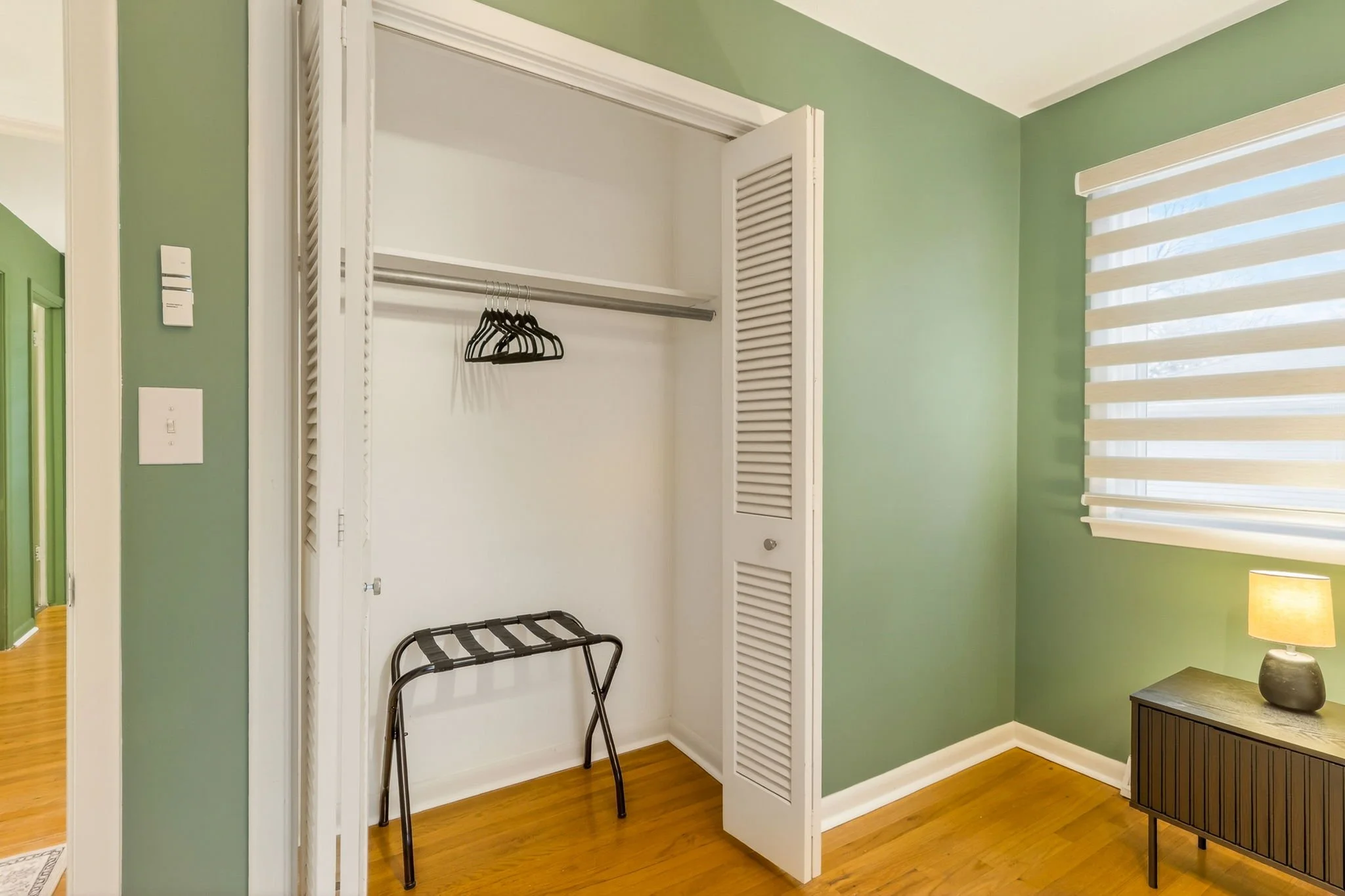 Empty closet with white doors, green walls, and hardwood floor, containing a few black hangers and a folded luggage rack. To the right, a window with white horizontal blinds and a small wooden side table with a lamp.