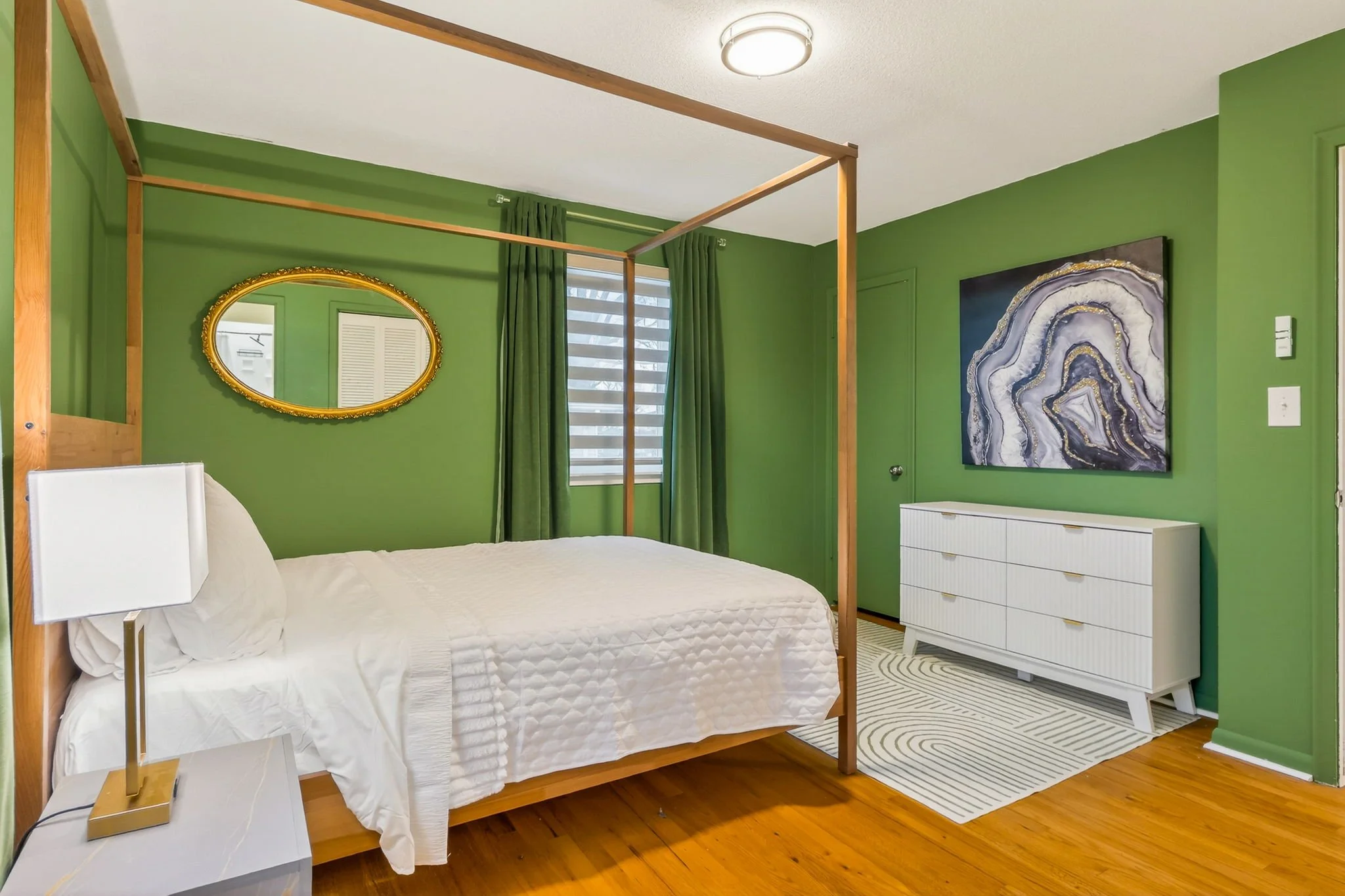Bedroom with green walls, a white bed, a wooden canopy frame, a white dresser, a decorative painting, an oval mirror, and a window with curtains.