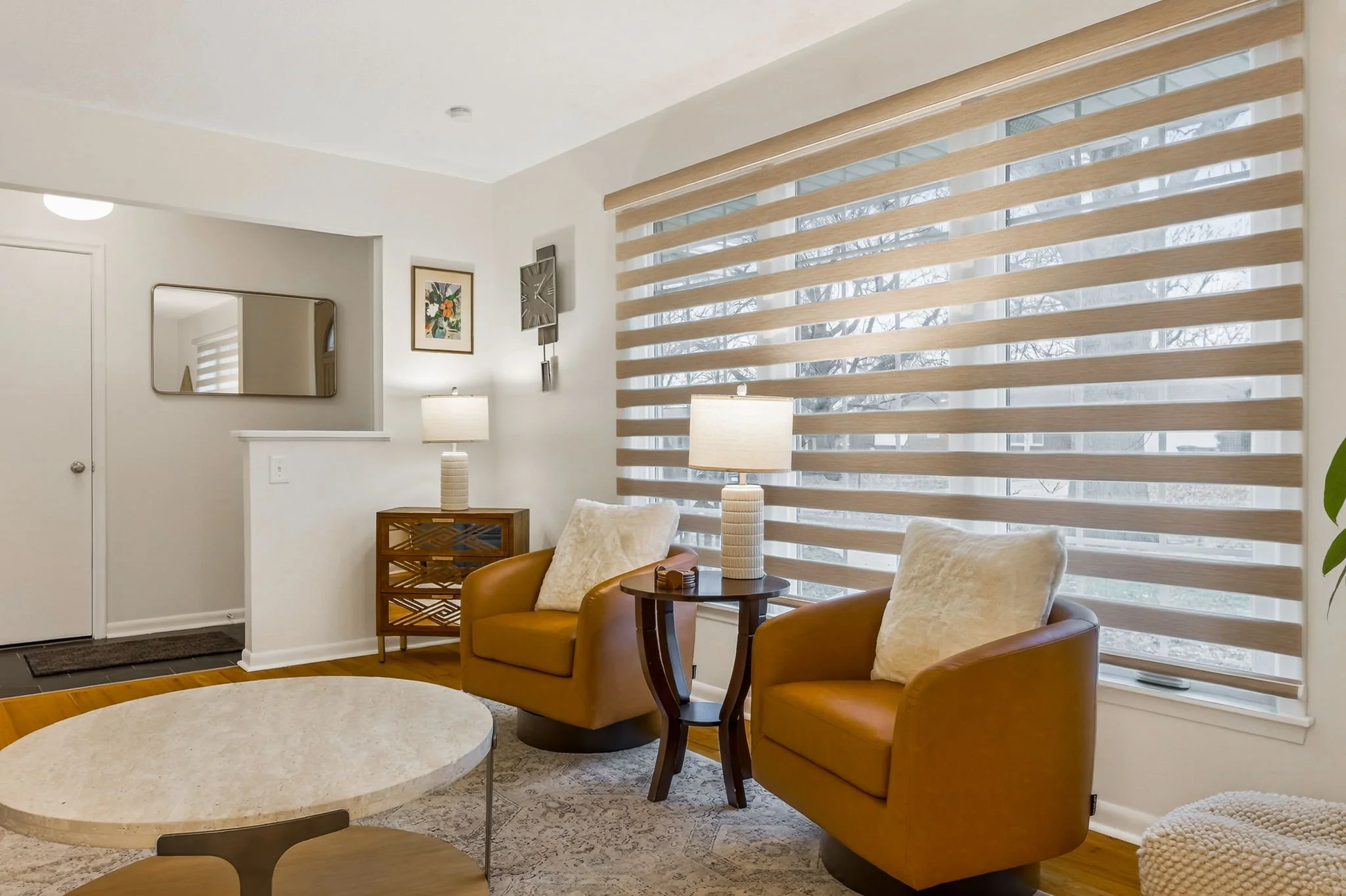 Living room with two tan armchairs, a small table between them with a lamp, a round coffee table, a mirrored wall decor, a wooden cabinet, and window blinds allowing natural light.