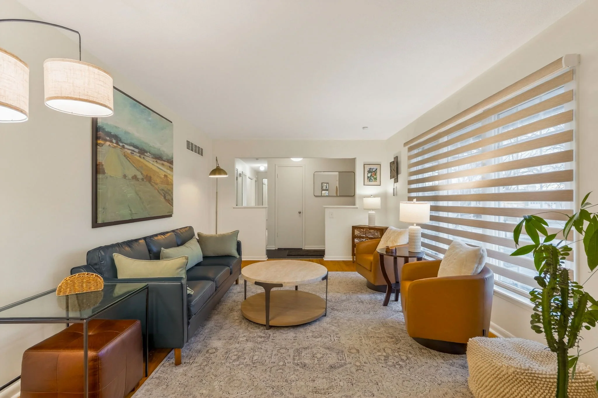 Living room with black leather sofa, yellow armchairs, wooden coffee table, side tables with lamps, framed artwork on the walls, patterned area rug, large window with striped blinds, and a potted plant.