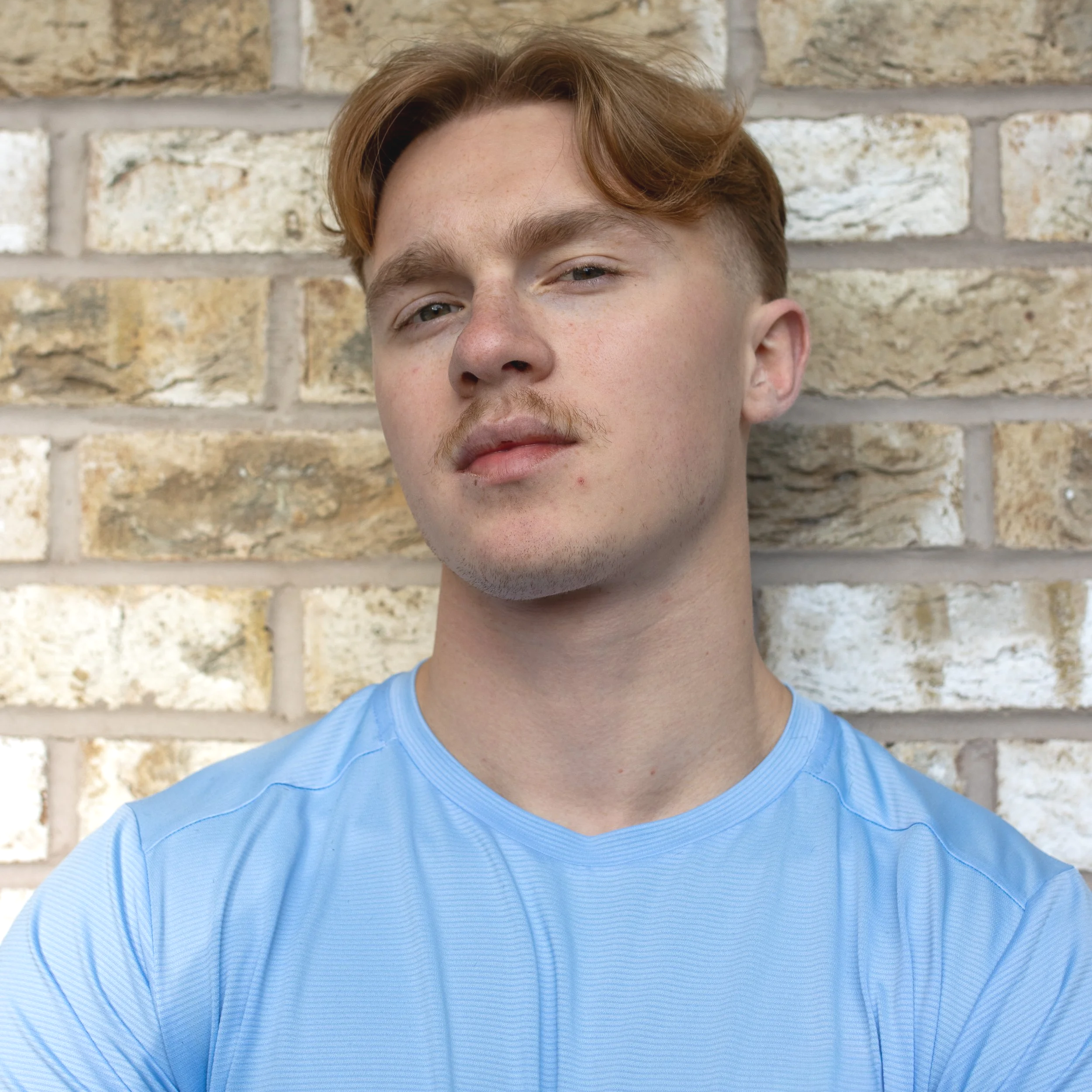 Close-up of a young person with red hair and facial hair, wearing a light blue shirt, standing in front of a brick wall.