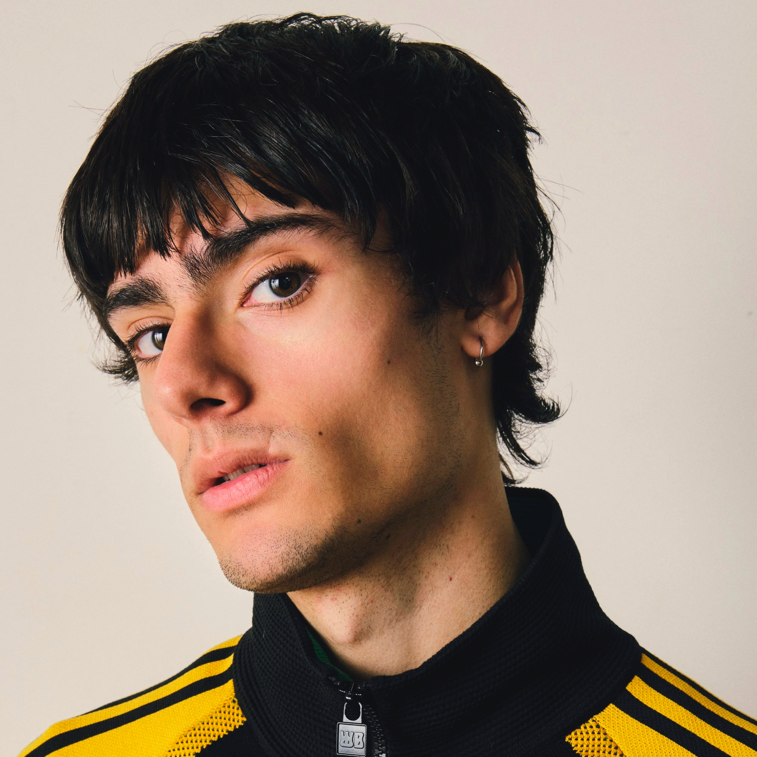 Close-up of a person with short black hair, brown eyes, and a small hoop earring in the left ear, wearing a black and yellow striped sports jacket with a zipper.