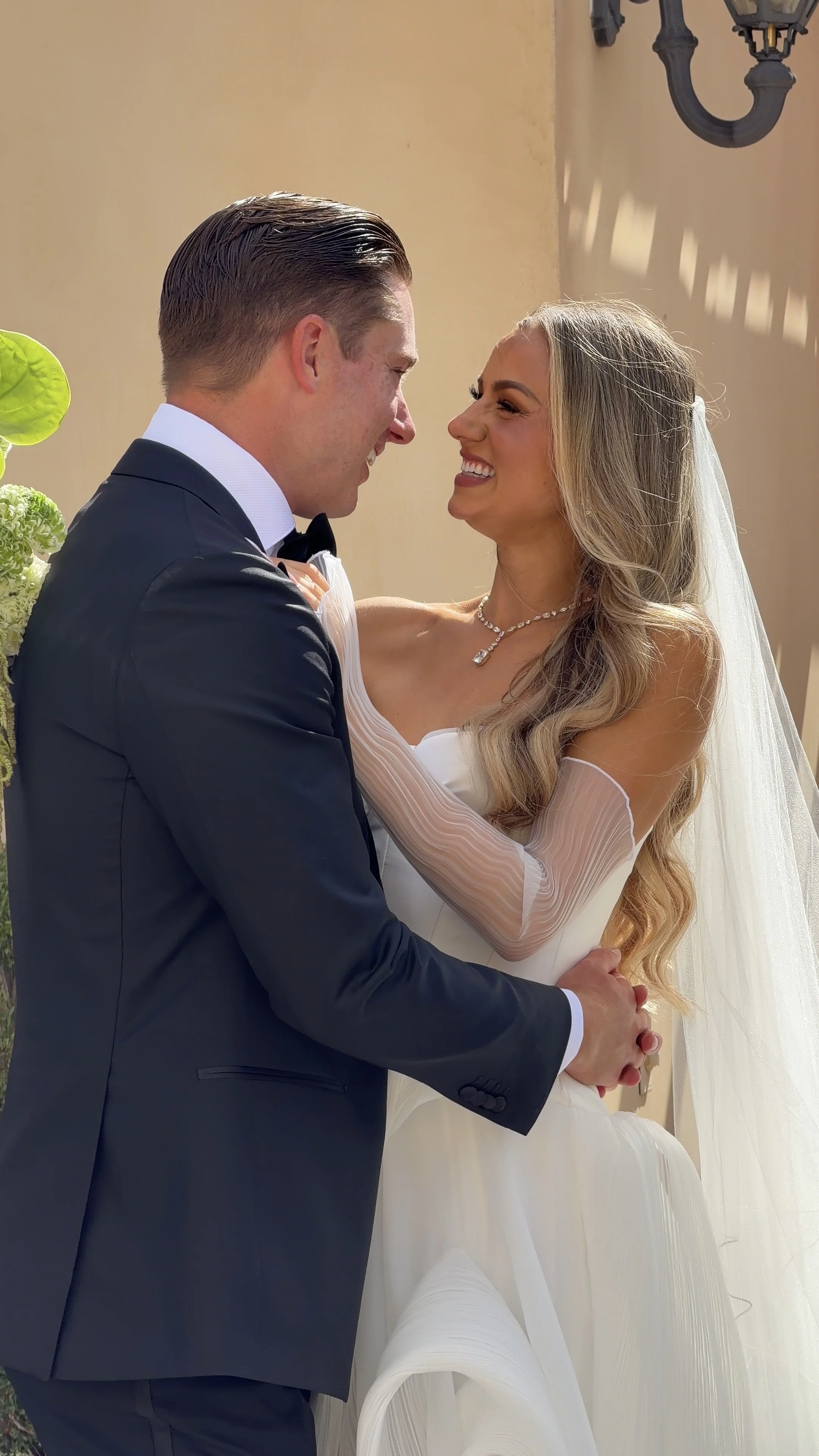 A newlywed couple is smiling and looking at each other, embracing during their wedding celebration, with a beige wall and a decorative outdoor light fixture in the background. AB Social Studio, Wedding Content Creator in California.