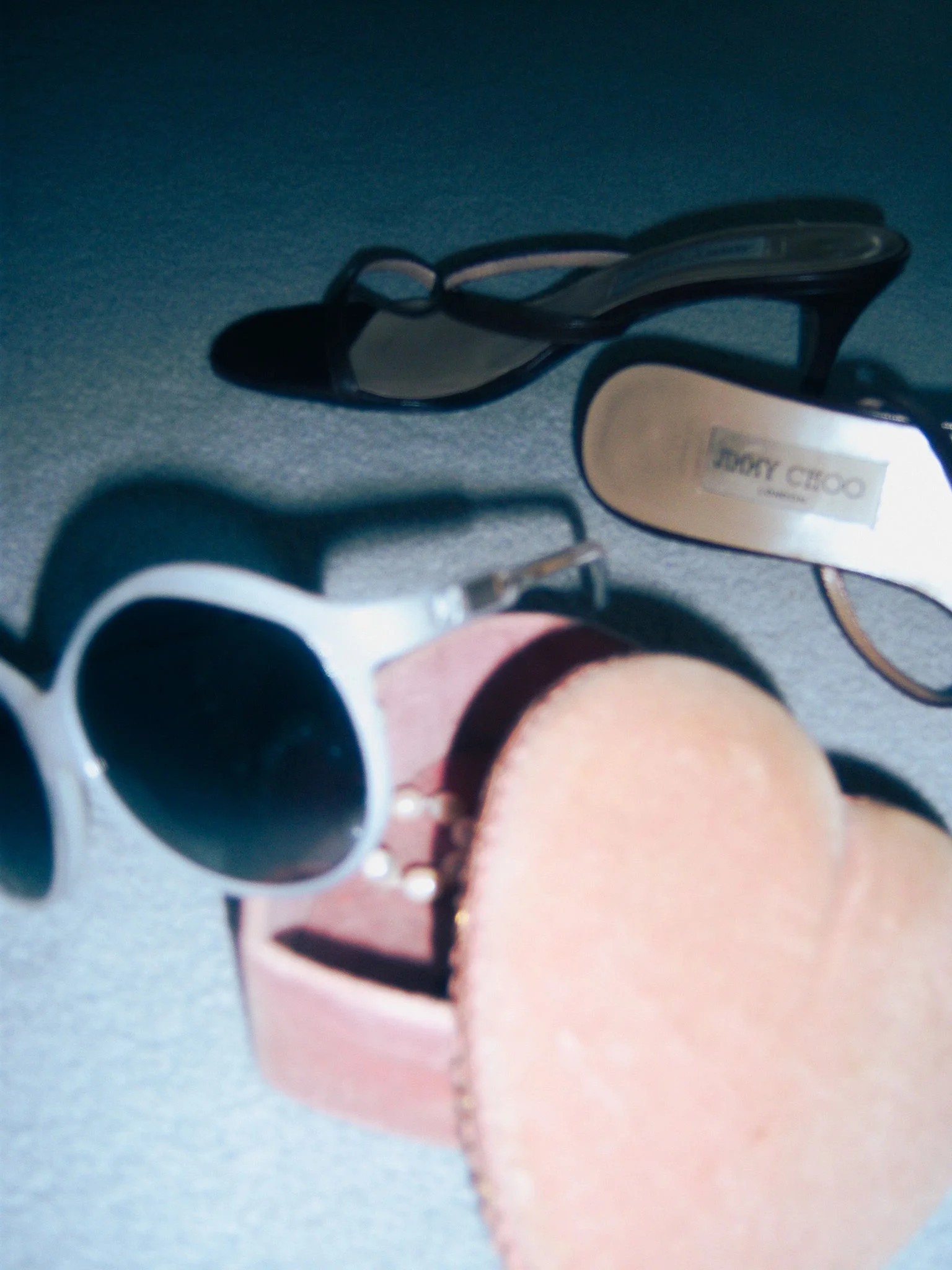 A pair of black high-heeled shoes and a pair of white sunglasses with black lenses resting on a gray surface, with a pink jewelry box and a beige suede object in the foreground.
