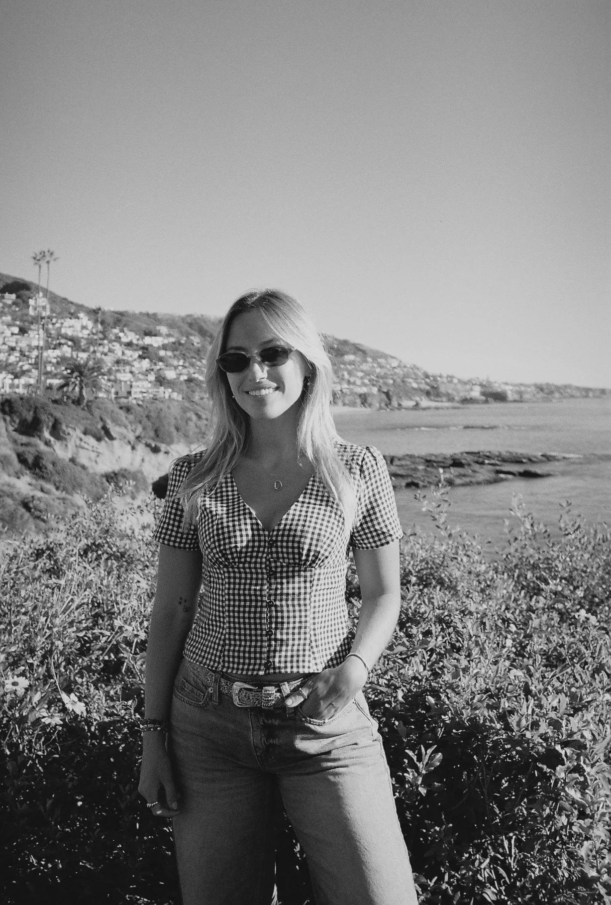 A woman standing outdoors near a coastal area with hills and houses in the background, wearing sunglasses, a checkered shirt, and jeans.