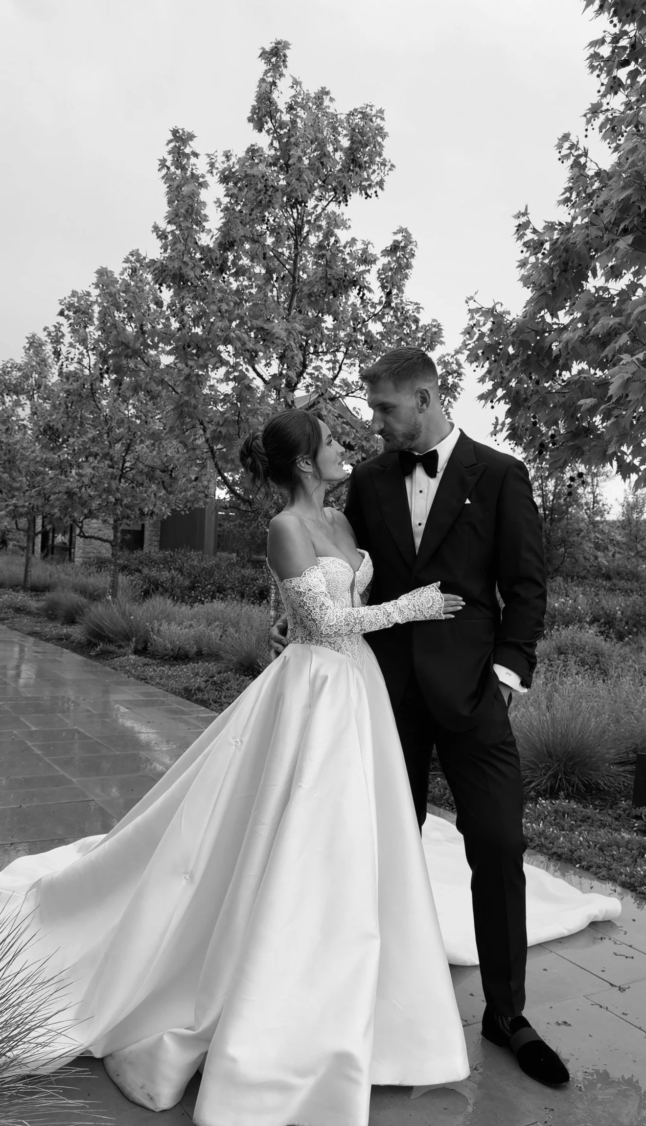 A black-and-white photo of a bride and groom standing closely outdoors, facing each other. The bride is in a strapless wedding gown with a long train, and the groom is in a tuxedo with a bow tie. They are surrounded by trees and garden plants.