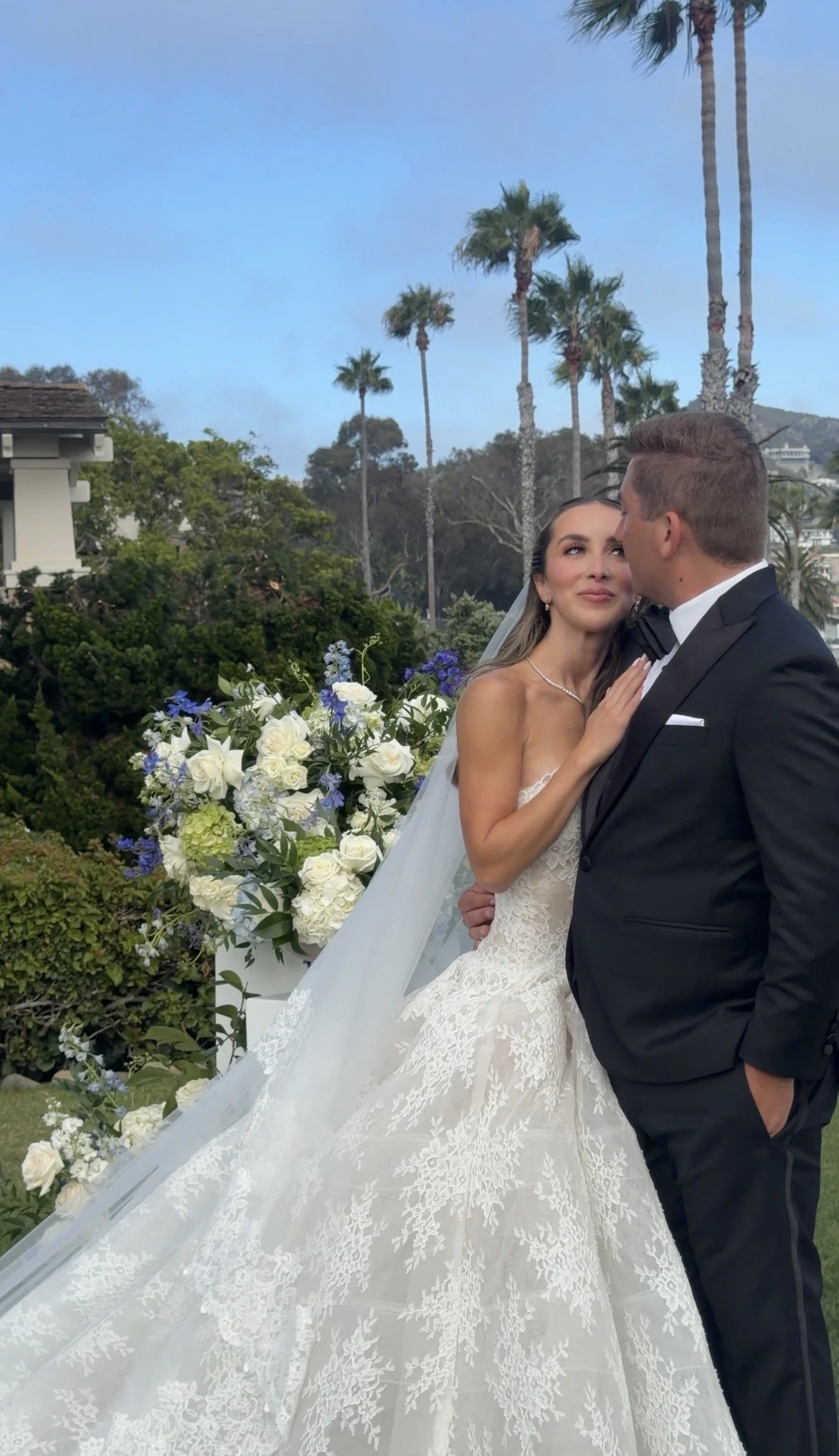A bride and groom are embracing outdoors at a wedding ceremony with palm trees, a large floral arrangement of white and purple flowers, and greenery in the background.