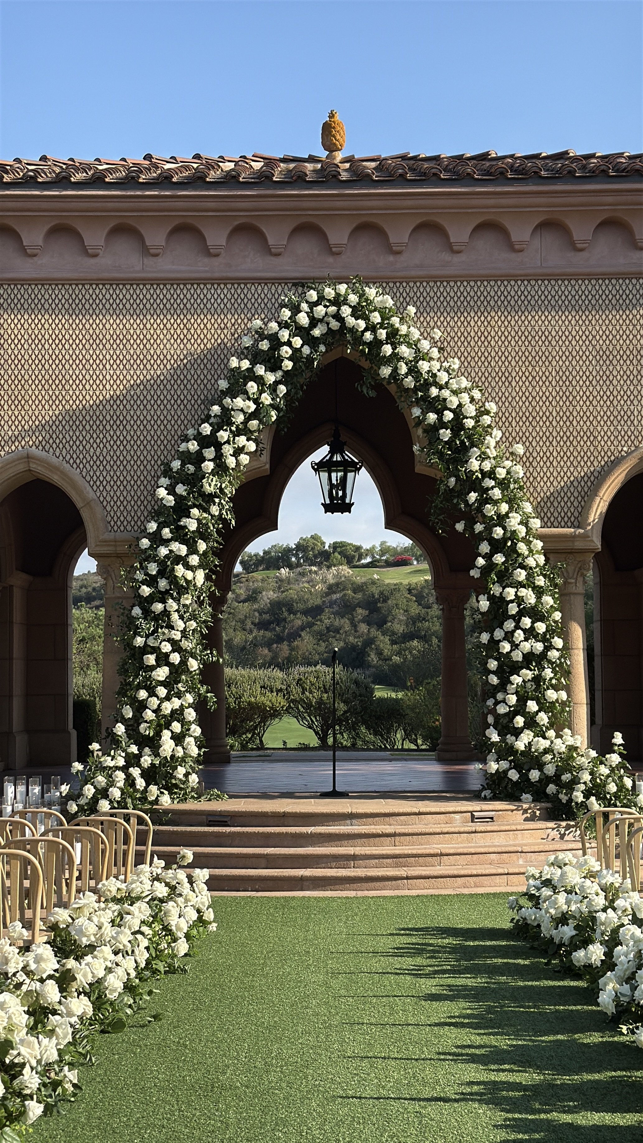 Outdoor wedding ceremony setup with a floral arch, chairs, and an aisle with green artificial grass under a clear blue sky. AB Social Studio, Wedding Content Creator in California.