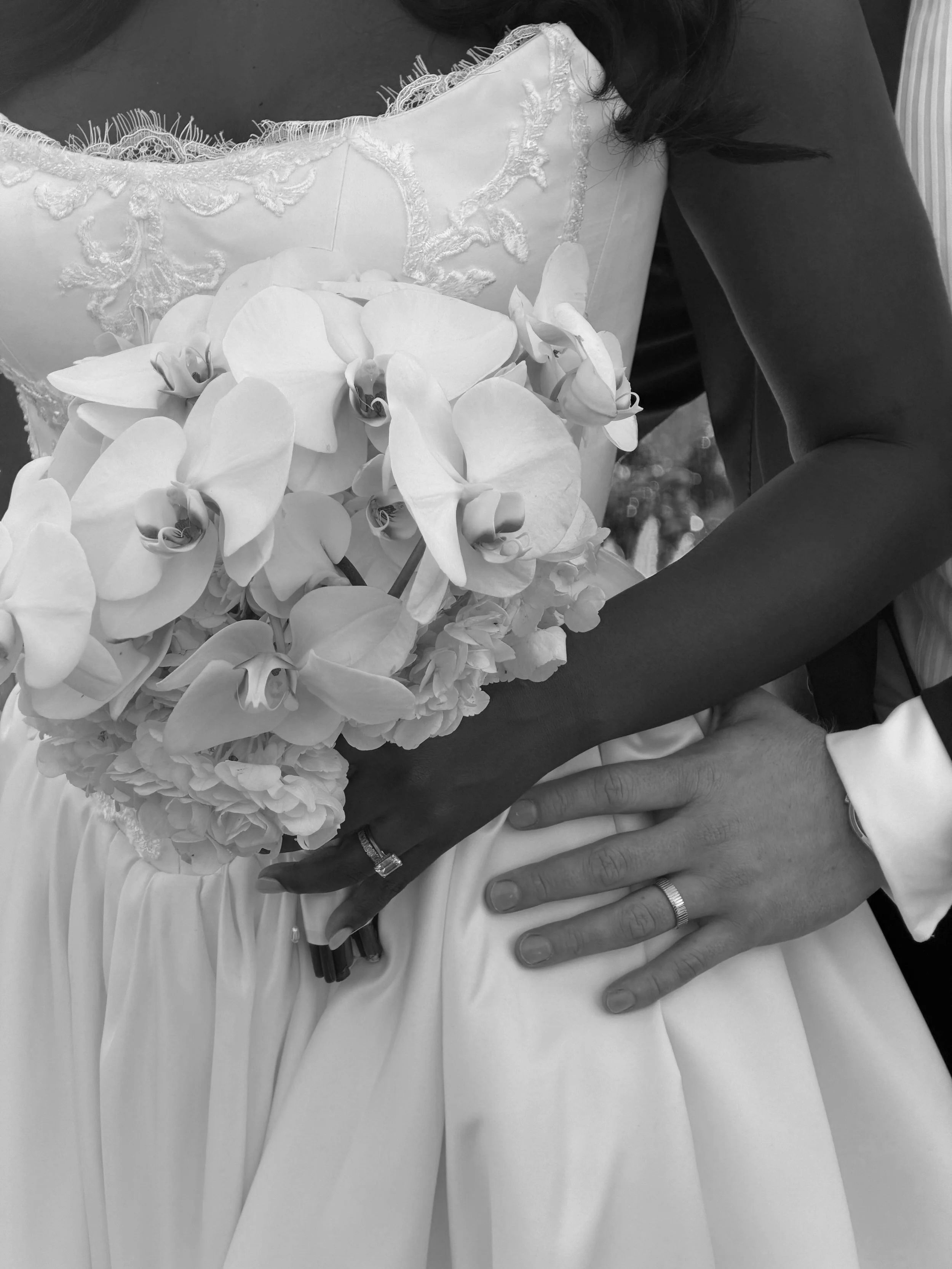 Close-up of a bride and groom's hands, with the bride holding a bouquet of orchids, on her wedding dress, both wearing wedding rings.