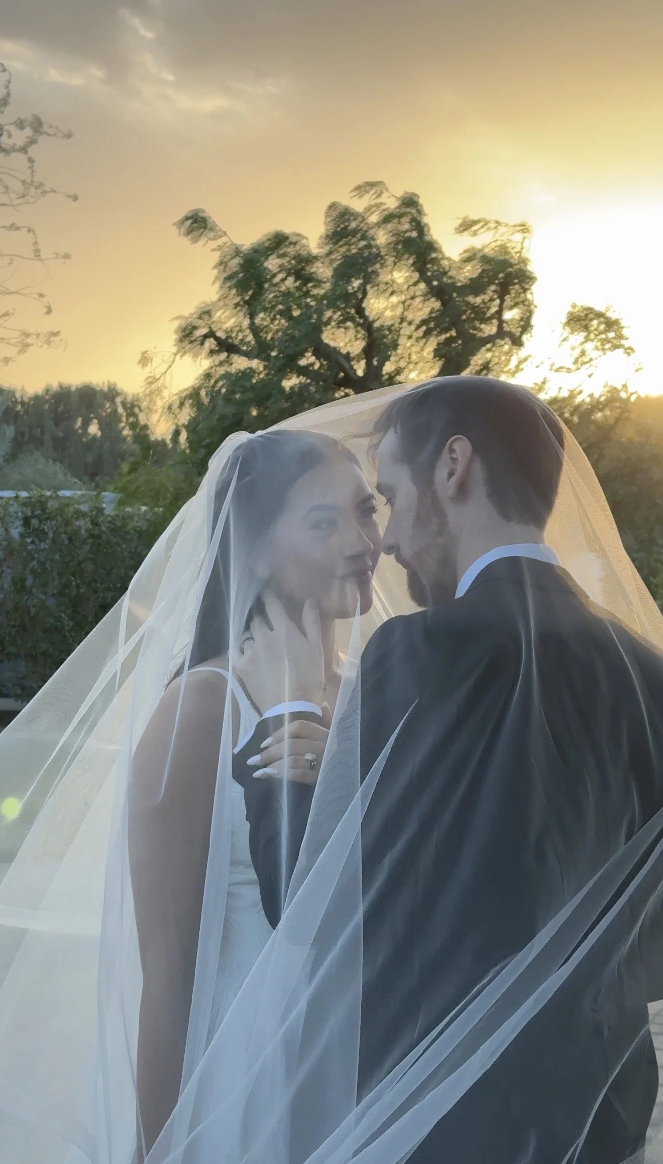 A bride and groom sharing a kiss outdoors at sunset, with the bride wearing a veil and the groom in a black tuxedo.