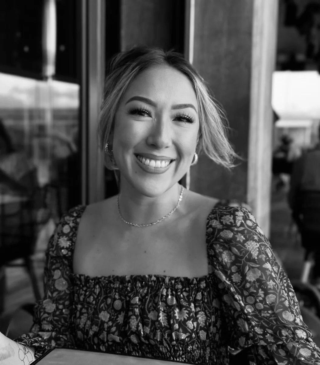 A woman smiling, wearing a floral dress, with earrings and a necklace, in a restaurant or cafe setting. AB Social Studio, Wedding Content Creator in California.