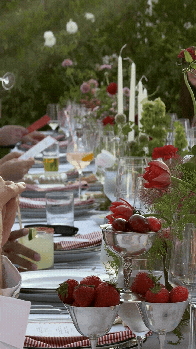 A long outdoor dining table decorated with flowers and candles, with people serving drinks and food.