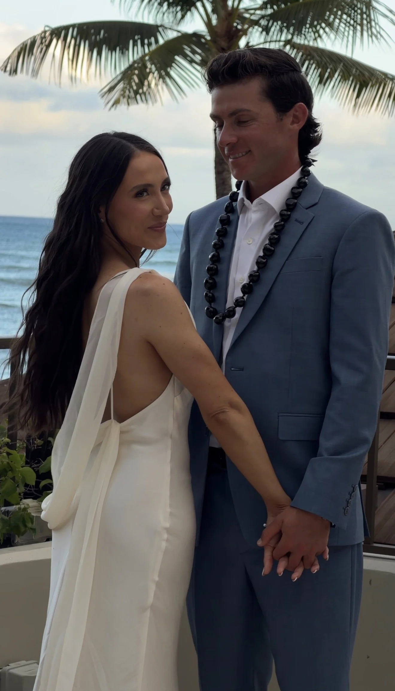 A couple holding hands on a beach with a palm tree behind them. The woman is wearing a white dress, and the man is in a blue suit with a white shirt and a black bead necklace. They are smiling and looking at each other.