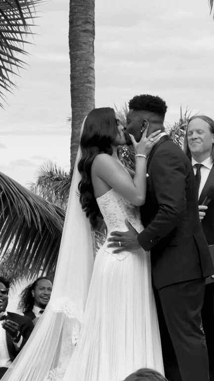 A bride and groom kiss during their wedding ceremony outdoors, with palm trees and friends in the background.