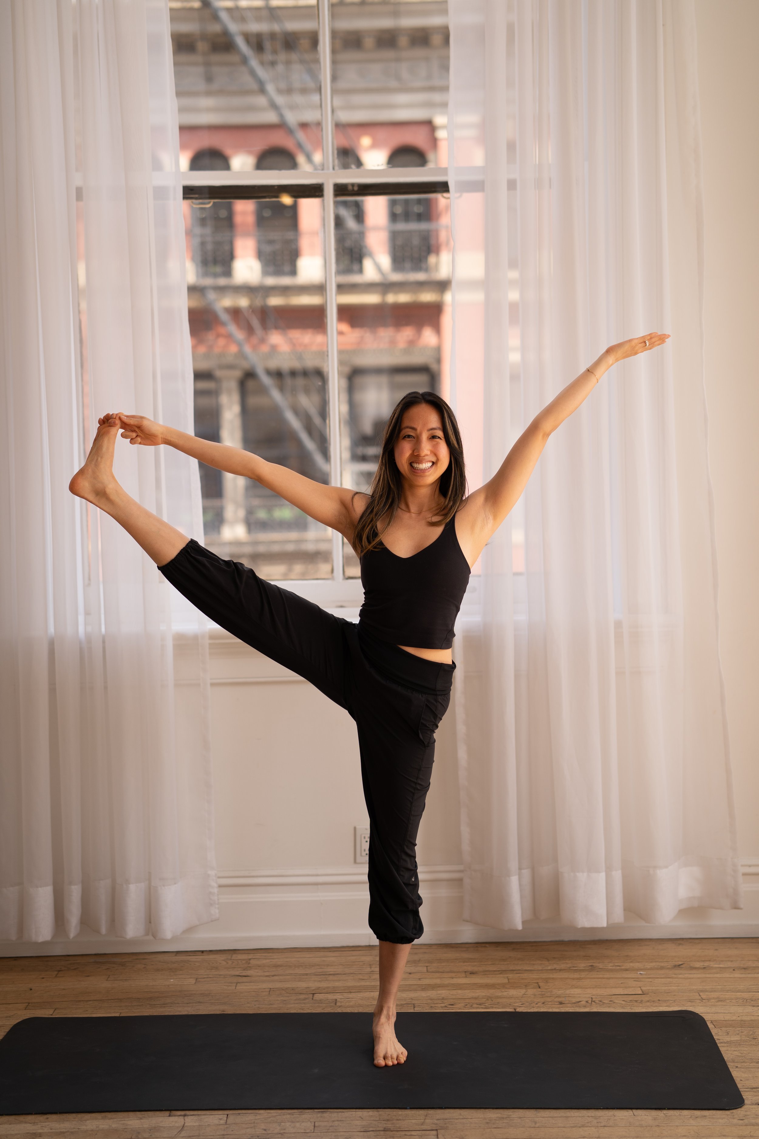 Woman practicing yoga, standing on one leg and holding the other leg with her hand in front, smiling near a large window with white curtains, urban buildings in the background.