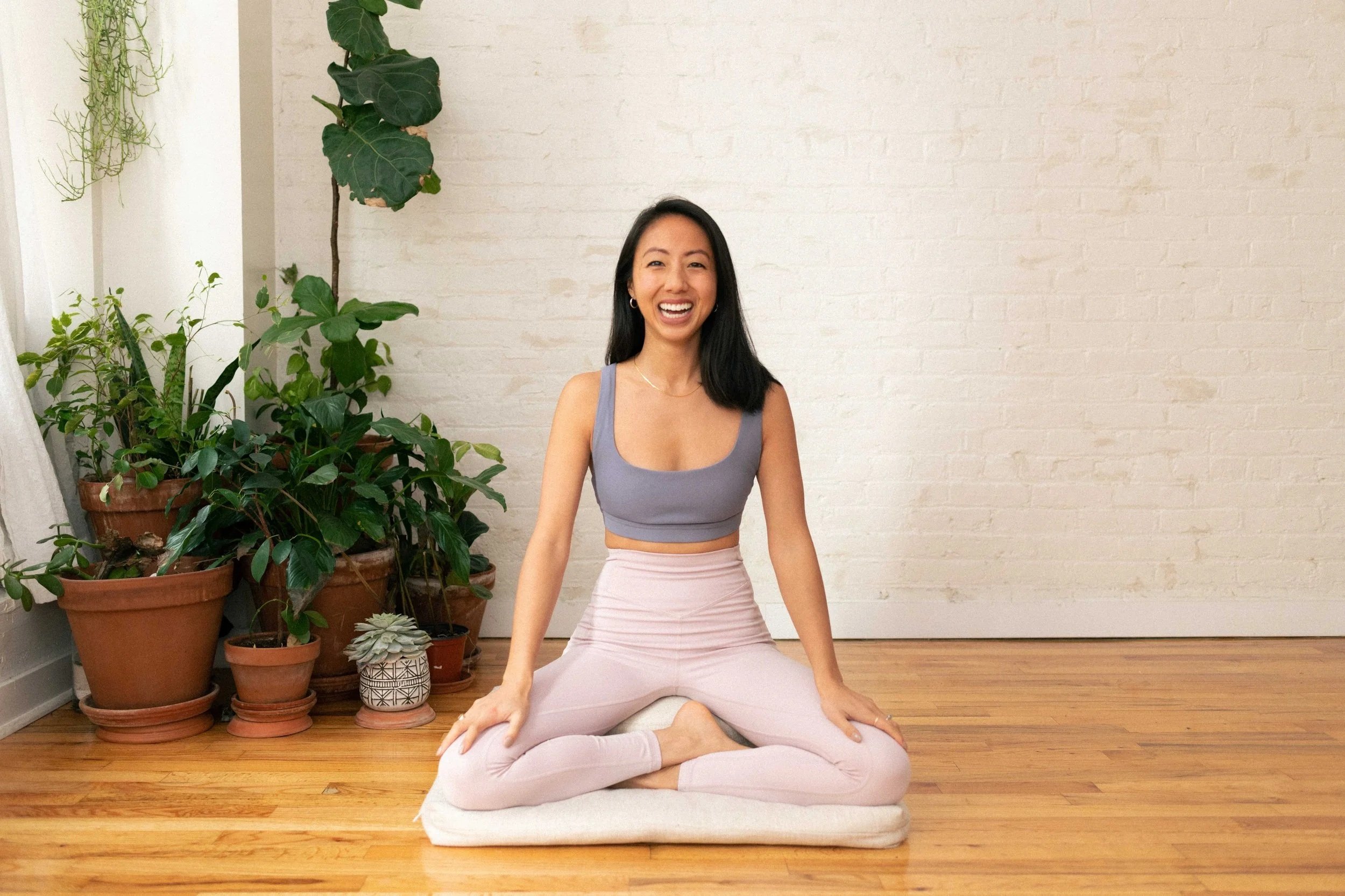 A woman sitting cross-legged on a cushion in a yoga pose, smiling in a bright room with potted plants and wooden flooring.