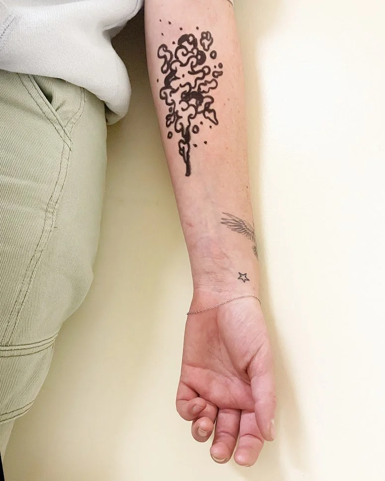 A person's forearm with three visible tattoos: a large black outline of a tree with leaves, a small star, and a wing with feathers. The person is wearing a white shirt and light-colored pants.