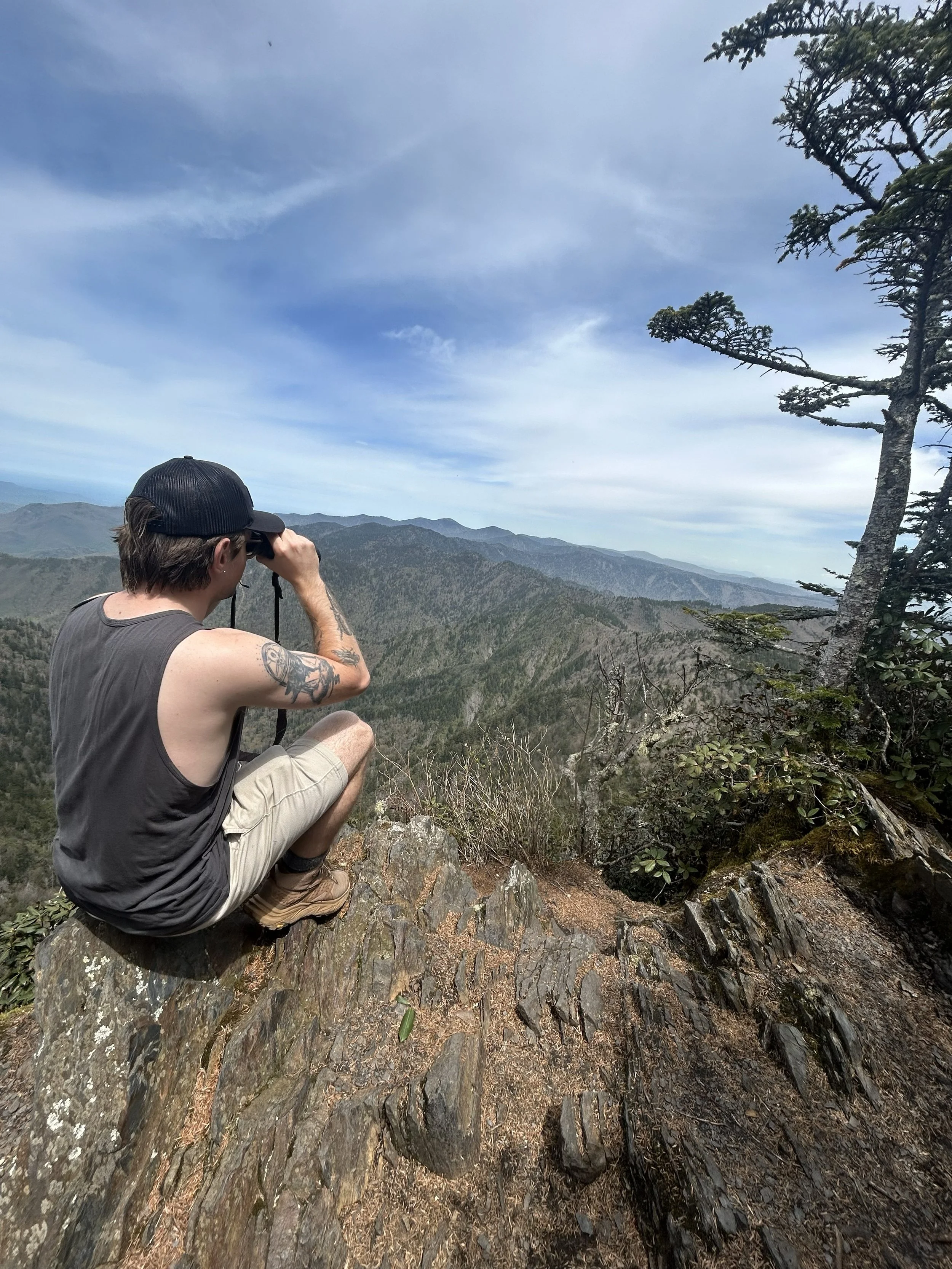 A man sits on a rocky outcrop taking a photograph of the distant mountains and sky with a camera. He has tattoos on his arm and is wearing a black cap, sleeveless shirt, and khaki shorts. There are trees and shrubs around him, and the sky is partly cloudy.
