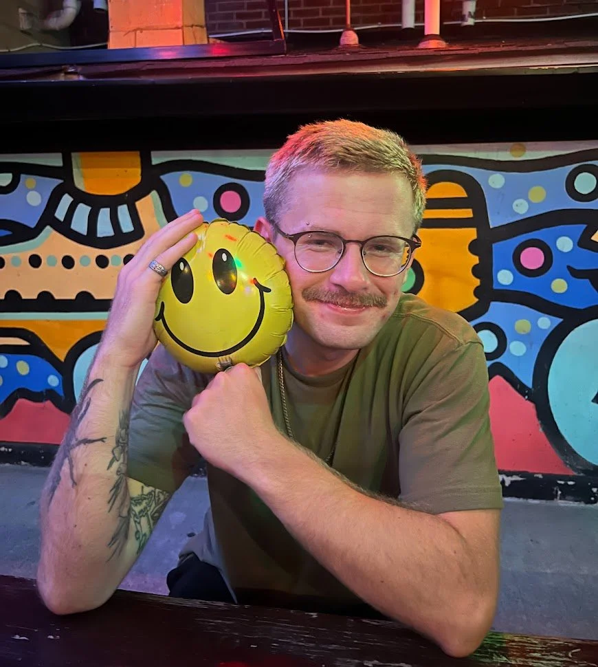 A smiling man with glasses, a mustache, and tattoos on his left arm is holding a yellow smiley face balloon close to his face. He is seated indoors in front of a colorful, abstract mural.