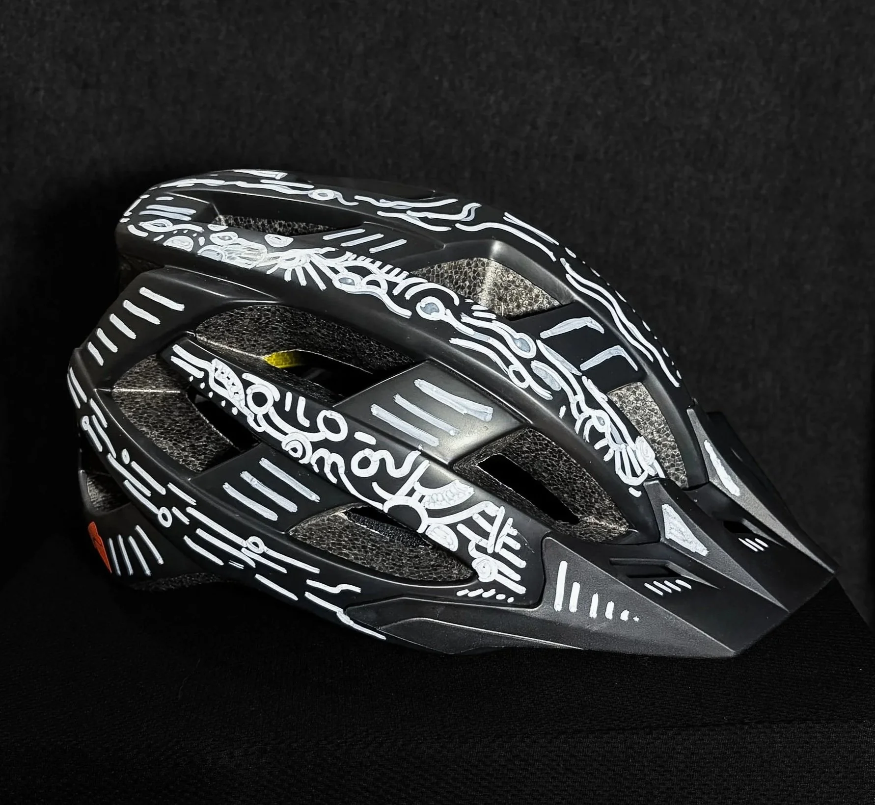 A black bicycle helmet with white graffiti-style design and patterns, placed on a black textured surface.