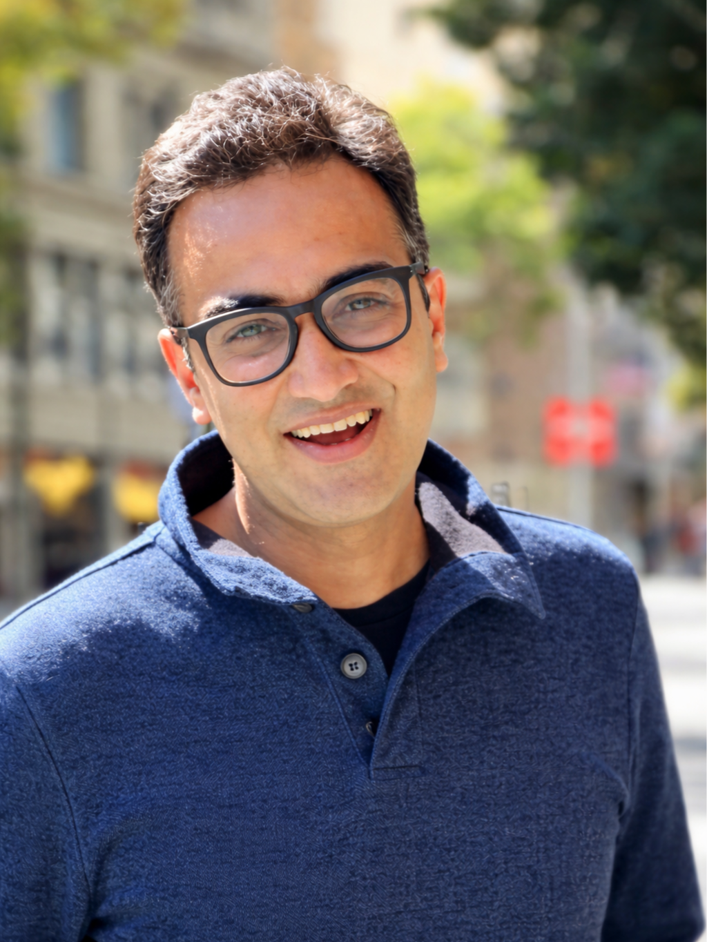 A man with glasses smiling outdoors on a sunny day, wearing a blue collared shirt with a patterned inner lining, with blurred buildings and trees in the background.