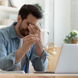 Mann sitzt vor einem Laptop mit Kopfhörern, sieht gestresst aus und hält sich die Stirn.