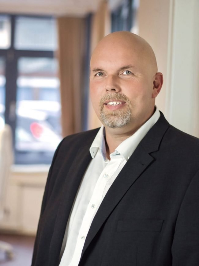 Ein Mann mit Glatze, Bart und Krawatte trägt einen schwarzen Anzug und steht in einem Raum mit Fenstern im Hintergrund.