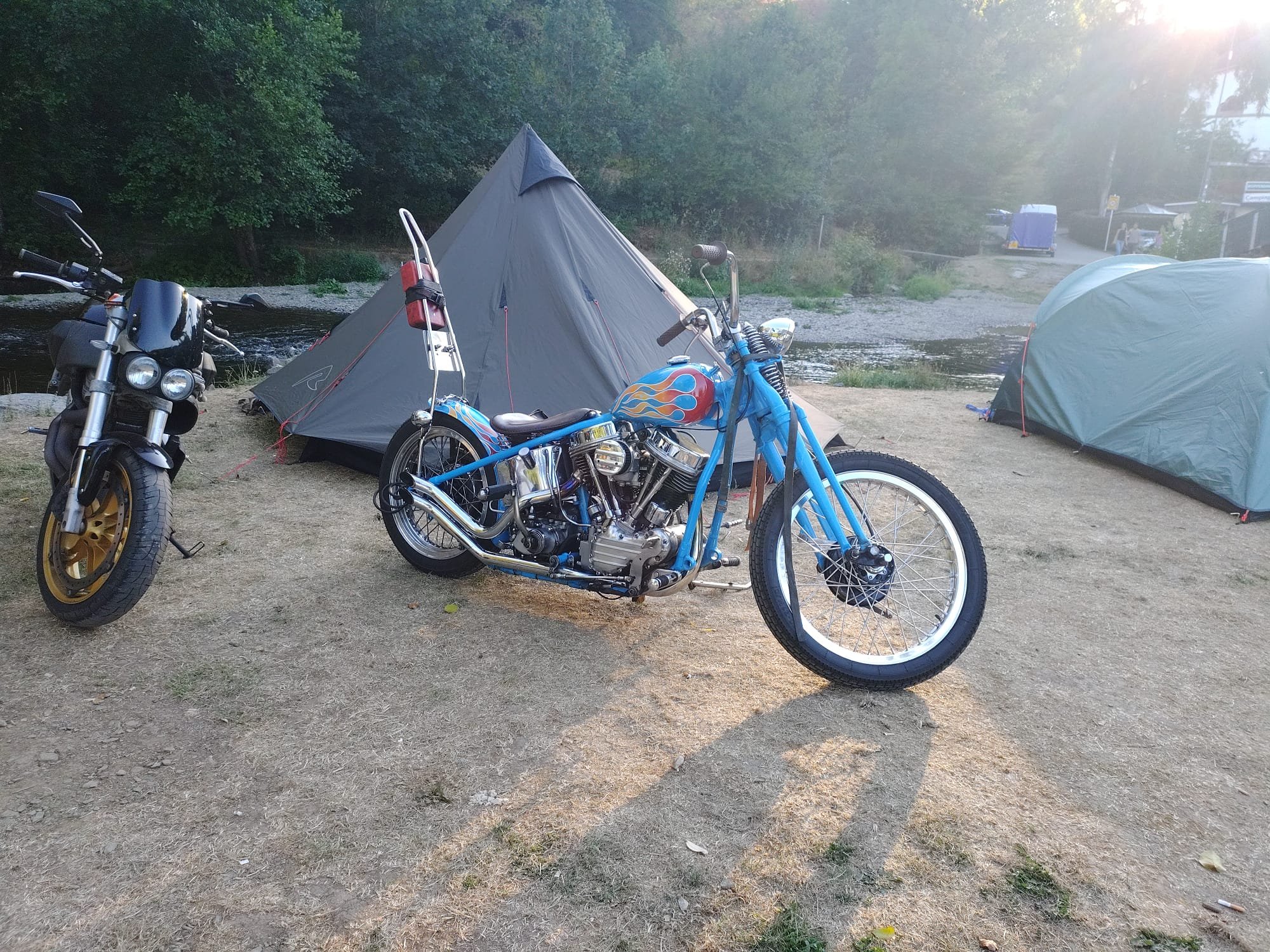 Een custom panhead op camping High Chapparal/Le Moulin, het zonlicht straalt door de bomen op de achtergrond.