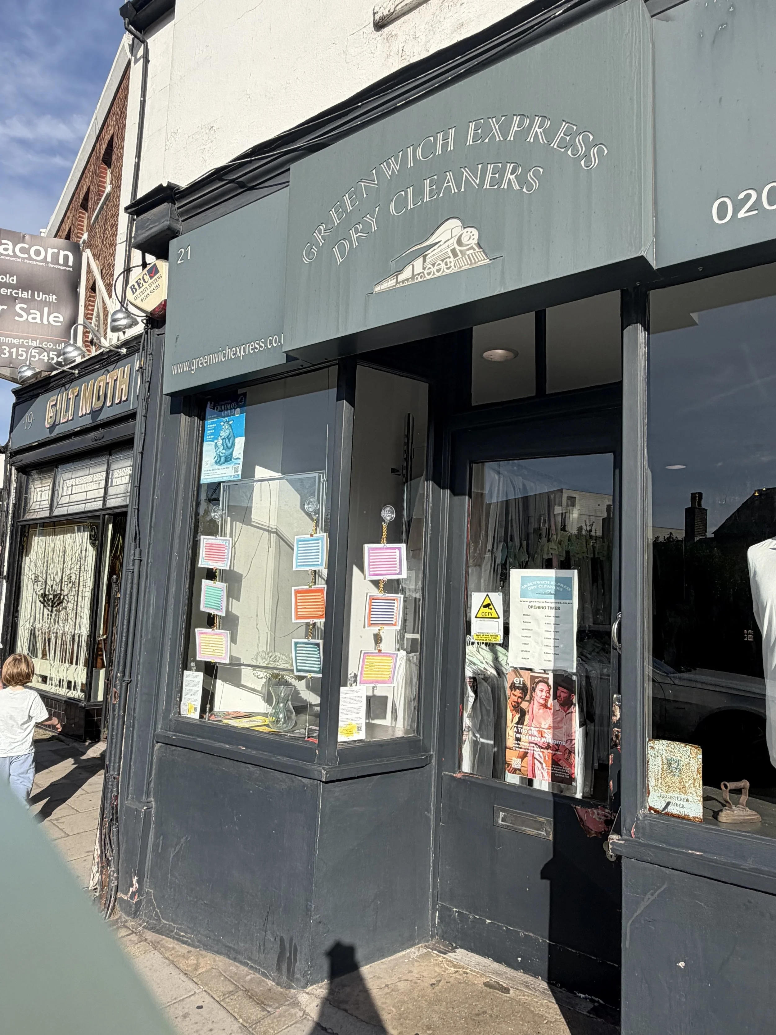 Front view of a storefront for Greenwich Express Dry Cleaners with a gray signboard, large glass window display, and black exterior walls. A small child is walking past the store on the sidewalk.