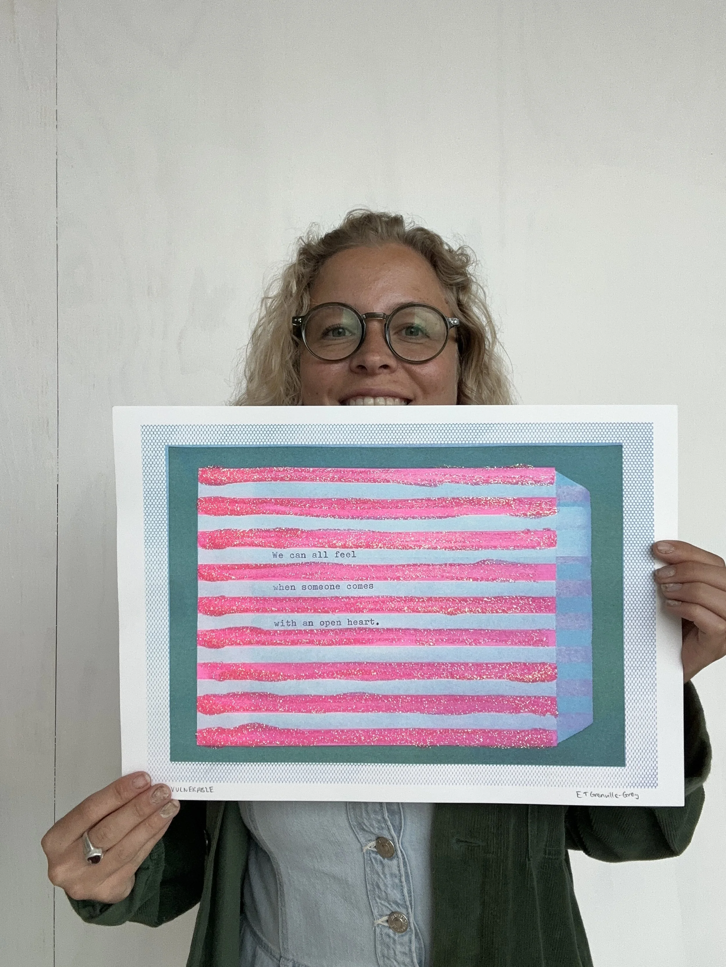 A woman with glasses and curly blonde hair holding a piece of artwork featuring pink and blue horizontal stripes with a quote about feeling with an open heart.