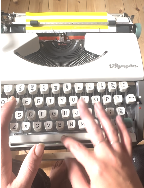 A white vintage typewriter with round gray keys and a gold brand logo, placed on a wooden surface, with hands approaching the keyboard.