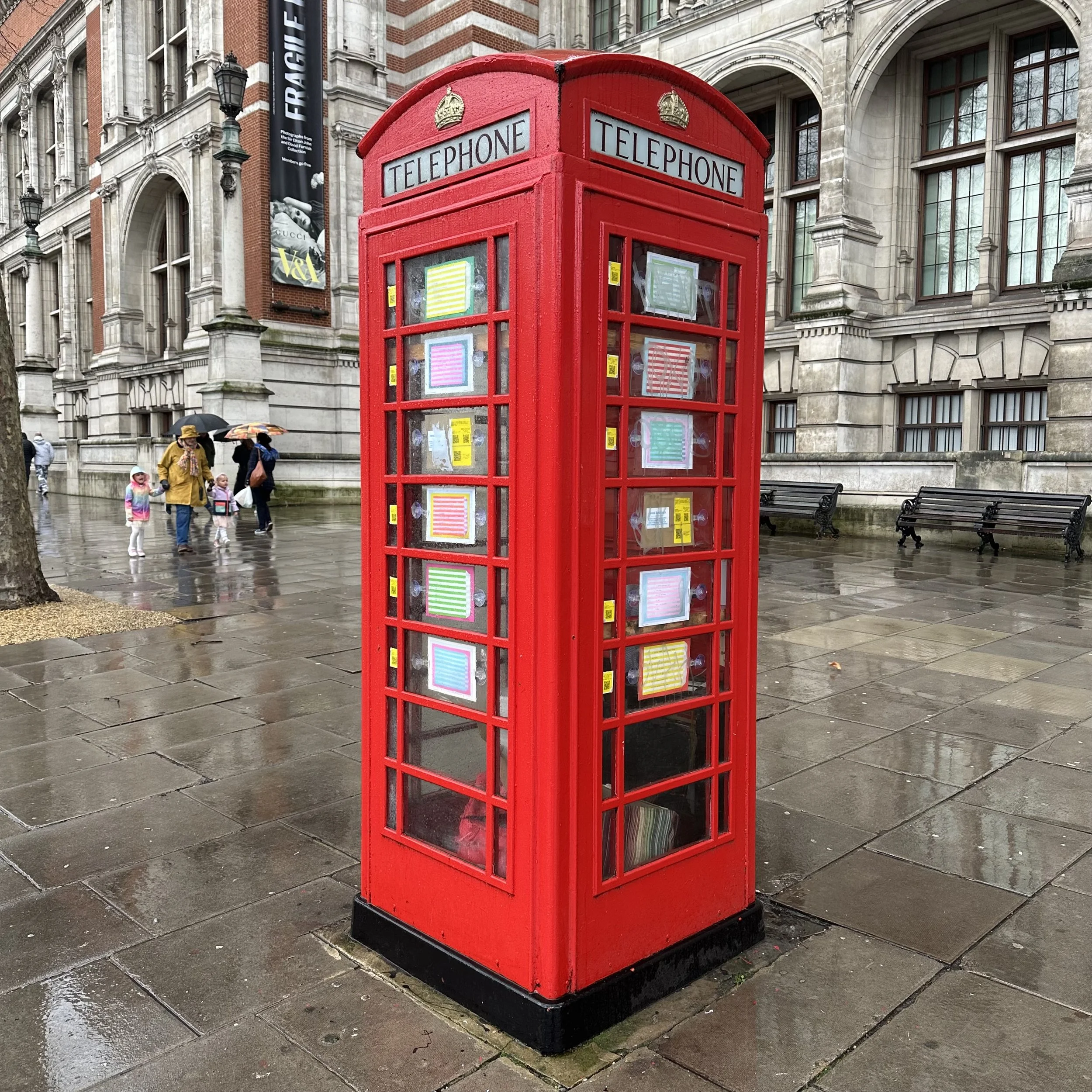 A traditional red British telephone booth on a wet city street with historic buildings in the background and pedestrians walking by.