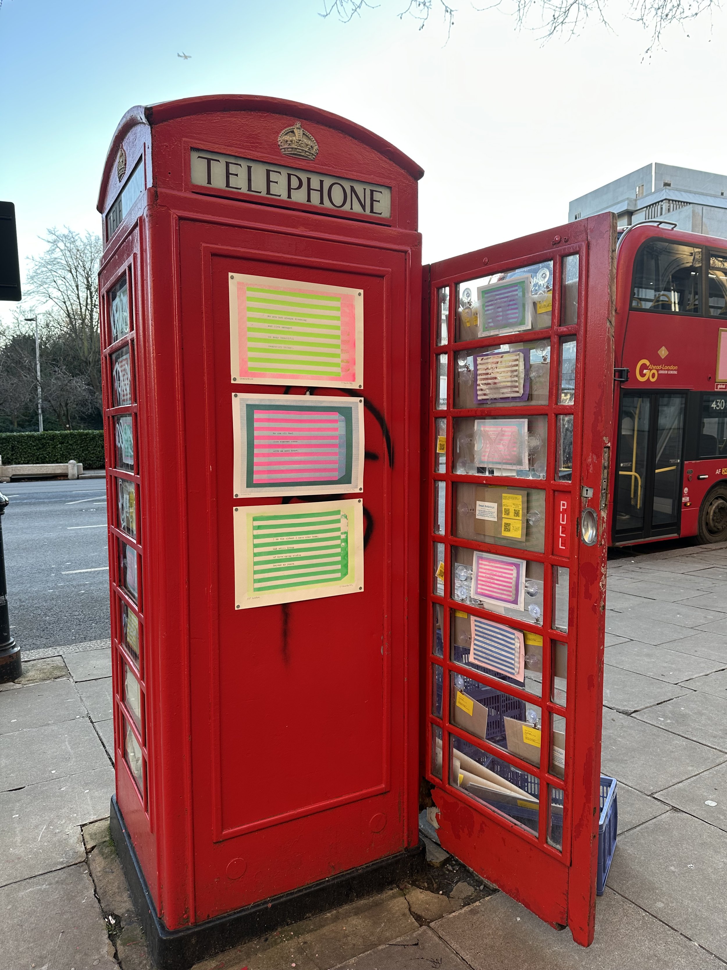A traditional red British telephone booth with its door open, revealing inside shelves with papers and small yellow objects, standing on a sidewalk near some parked cars and a city street.