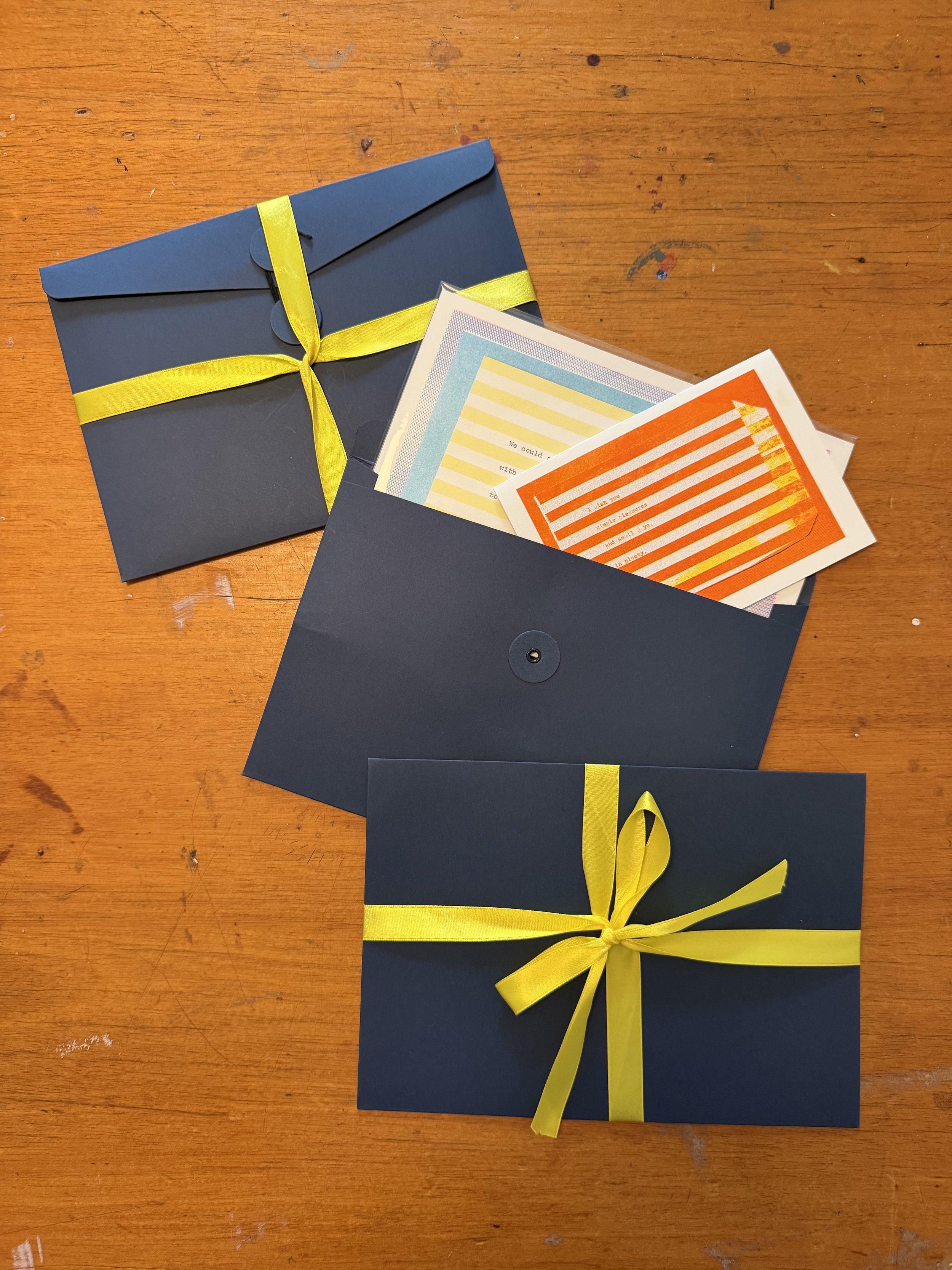 A dark blue envelope decorated with a yellow ribbon bow, placed on a wooden surface. Inside the envelope are colorful cards with striped patterns in yellow, blue, and orange.