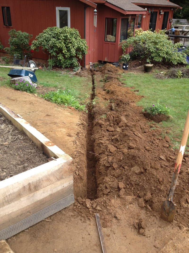 A trench being dug in a backyard garden near a red house, with a shovel resting nearby and a partially built raised garden bed on the left. There are plants, bushes, and small trees around the area.