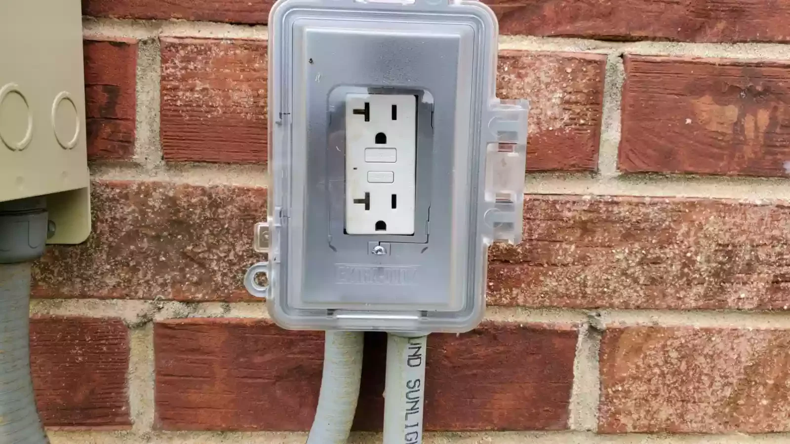 An outdoor electrical outlet mounted on a red brick wall, with a clear plastic weatherproof cover and a gray conduit running down from the bottom.