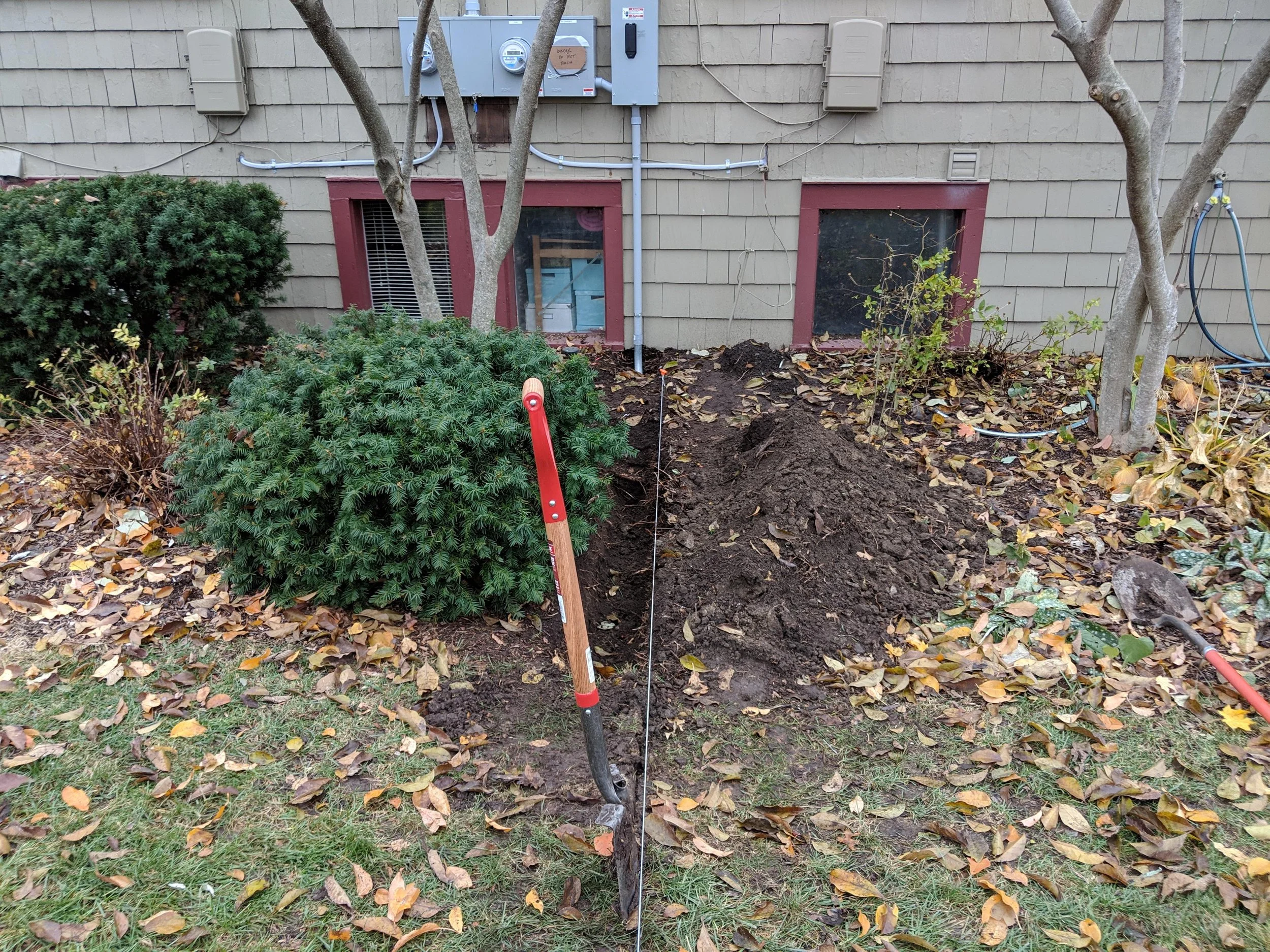 A gardening project with a small pile of dirt, a handheld garden tool, and a string line marking a gardening bed next to bushes and trees outside a house.