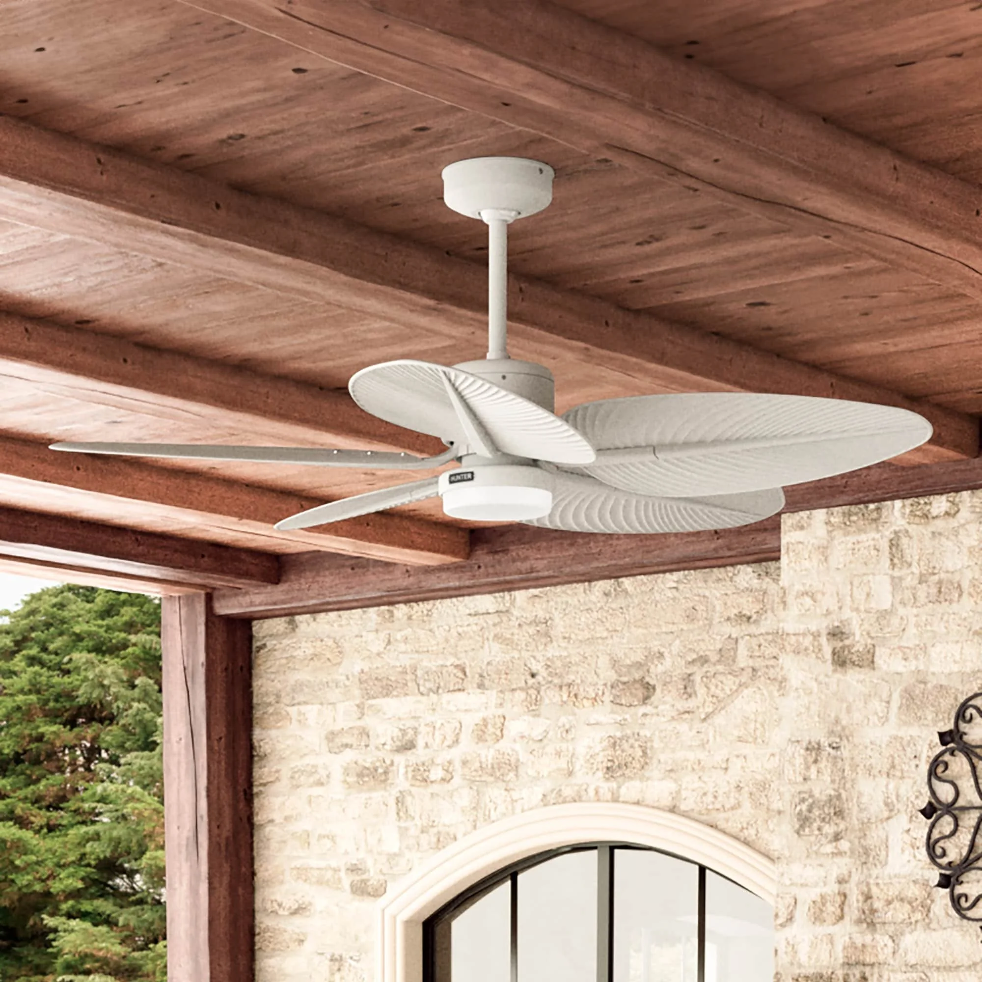 White ceiling fan with palm leaf-shaped blades mounted on a wooden ceiling in an outdoor patio.