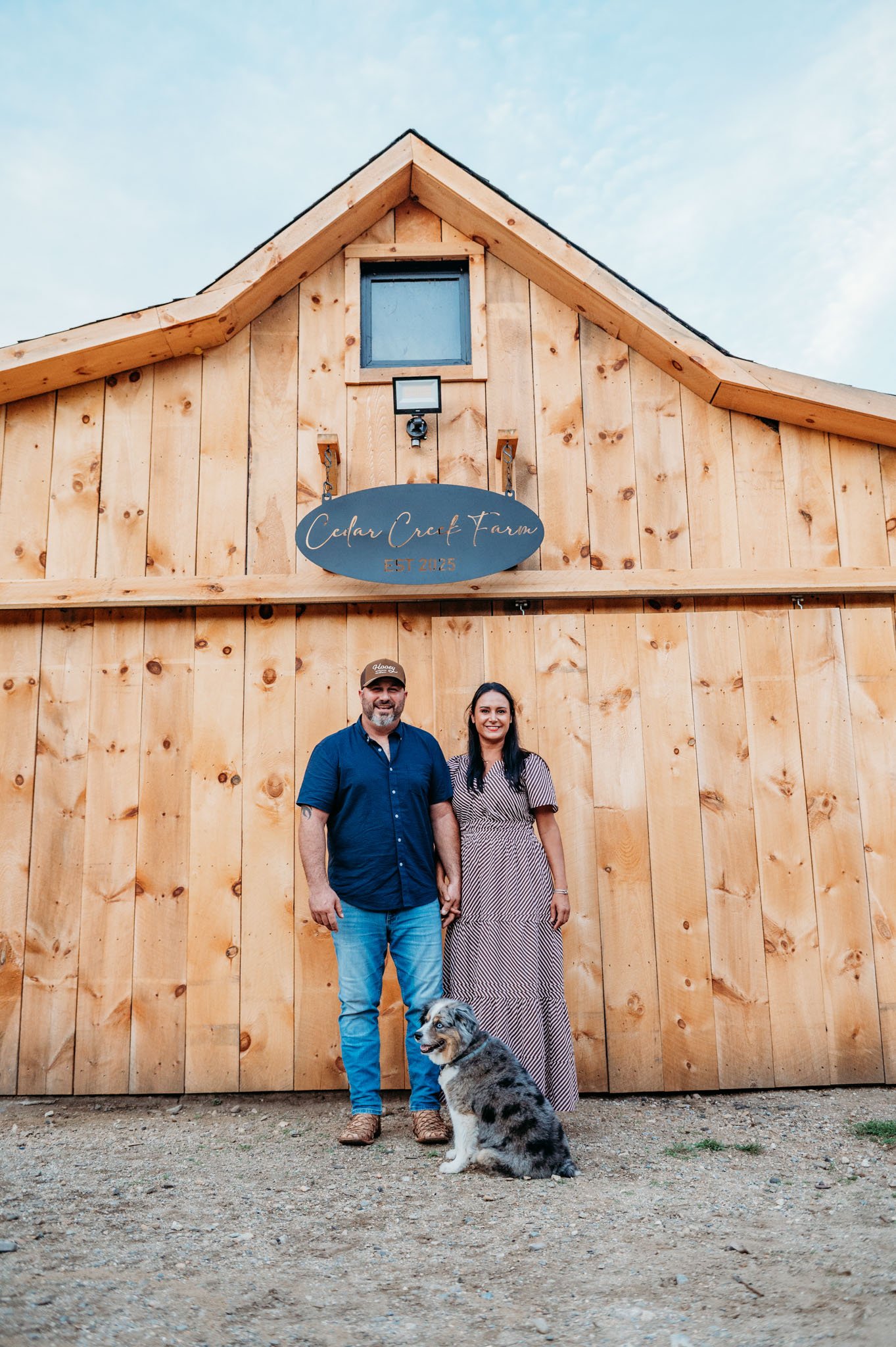 Ashley and rob and Australian Shepherd, Covey, stand outside Cedar Creek Farm, a wooden barn with a sign, under a clear sky at cedar creek farm connecticut dog breeder puppies for sale near me 